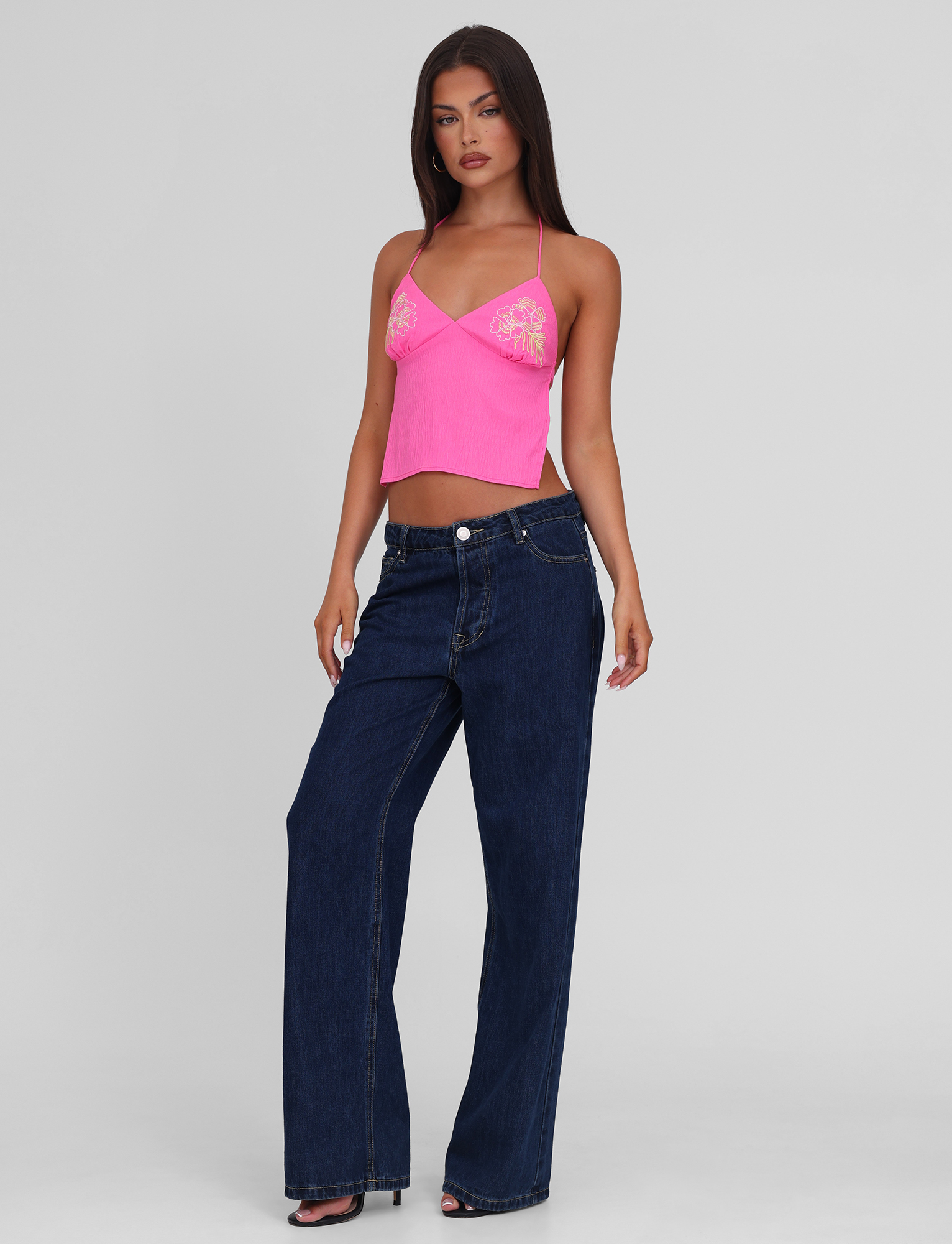 A woman poses against a plain background wearing the TIGERMIST KAELAH TOP in hot pink, a halter style with lace detailing, paired with loose-fitting dark blue jeans. She has long straight hair and poses with one hand by her side and the other slightly bent.