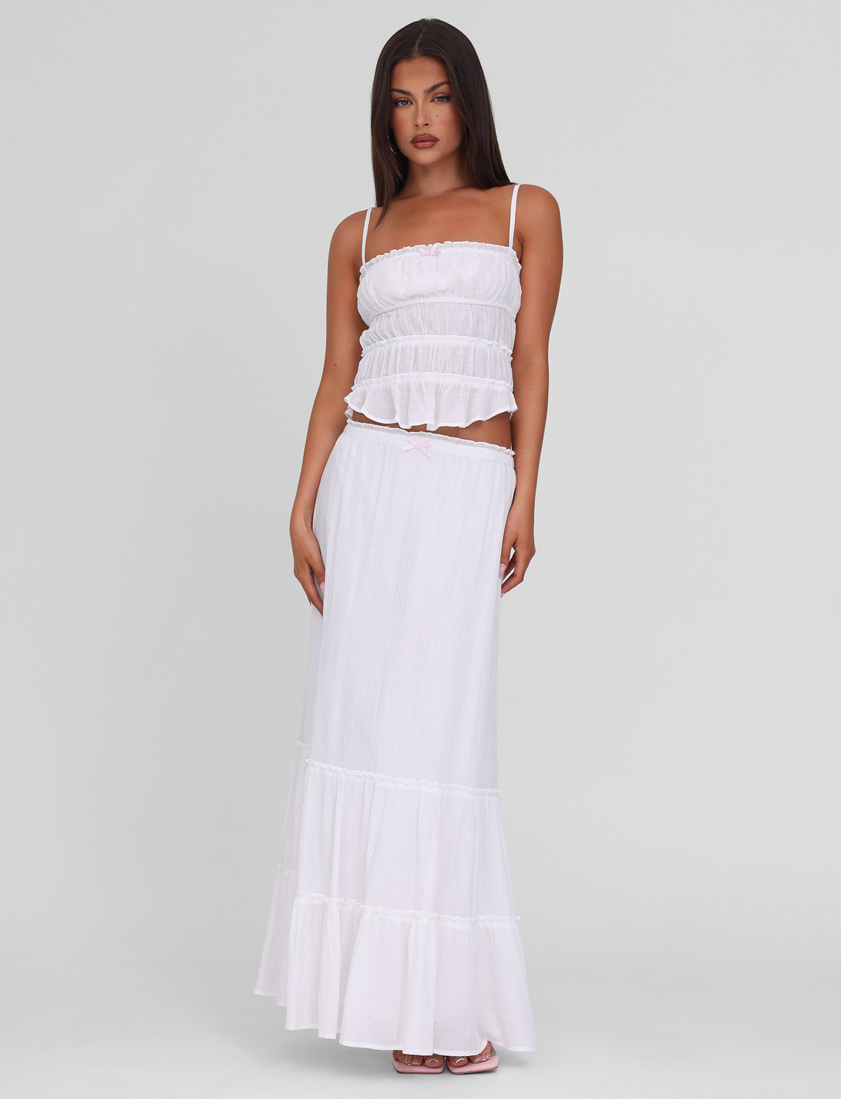 A woman with long dark hair and a neutral expression stands against a plain background wearing the TIGERMIST ASHLYNN TOP in white, featuring adjustable shoulder straps and tiered detailing. One hand rests by her side as she pairs it with a flowing white skirt.