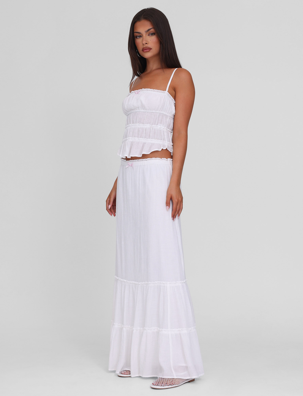 A woman with long dark hair models the TIGERMIST ASHLYNN MAXI SKIRT in white, featuring a tiered design and elasticated waist. She wears it with a sleeveless crop top, standing in profile against a plain light background.