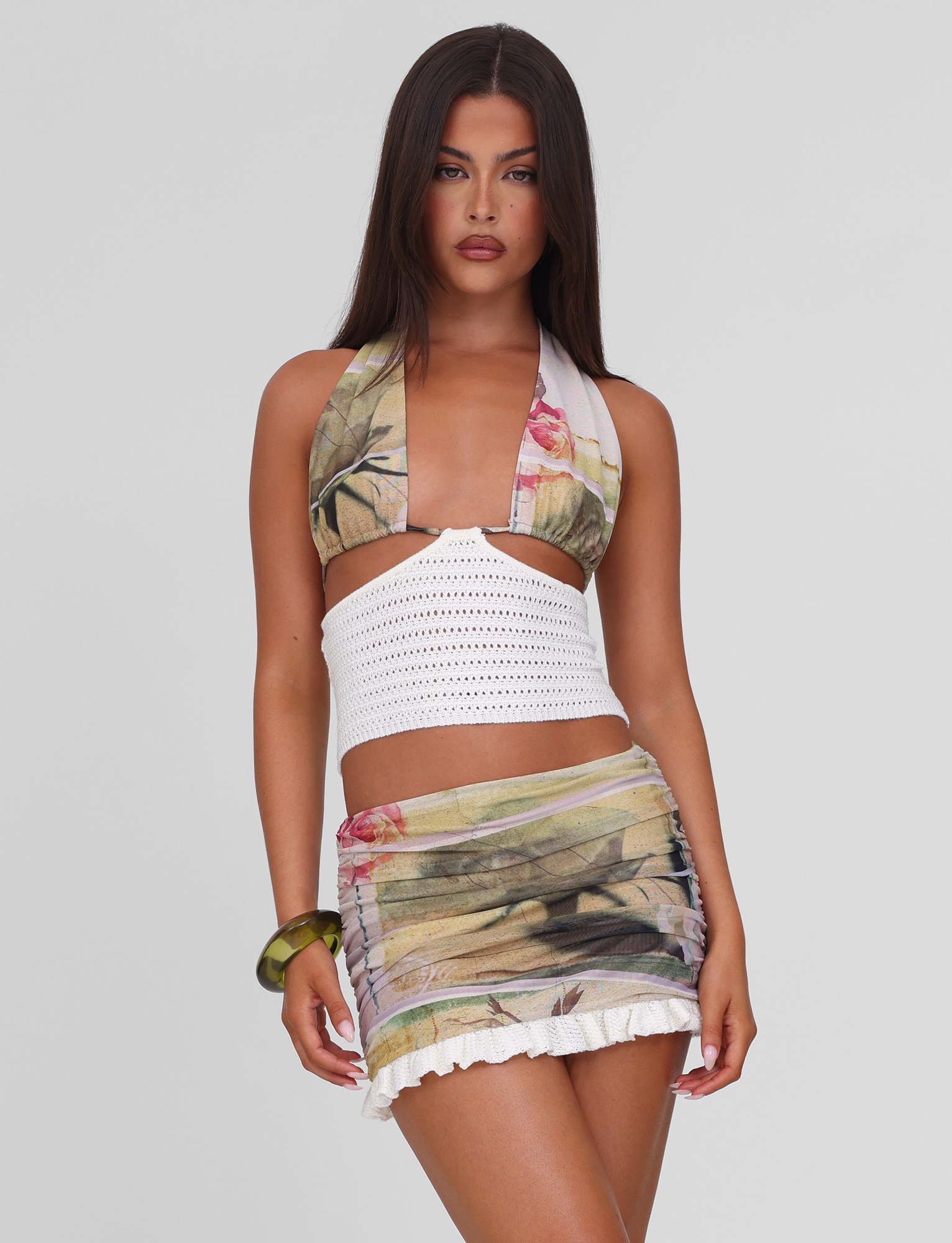 A woman with long dark hair wears the TIGERMIST TILLEY MINI SKIRT - ROSE POSTCARD, a floral mesh mini skirt with an exclusive print and white ruffled hem, paired with a floral halter crop top. She stands against a plain light background.