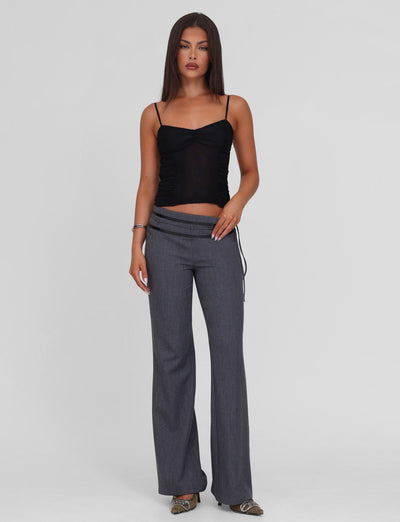 A woman with straight, long brown hair wears the TIGERMIST KALLIE TOP - BLACK, a lace camisole with adjustable straps, paired with gray low-rise wide-leg pants featuring a slightly asymmetrical waistband and open-toed shoes.