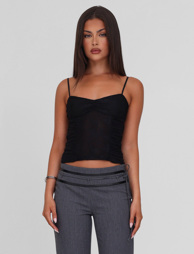 A woman with straight brown hair wears the TIGERMIST KALLIE TOP in black, a fitted lace camisole with adjustable straps, paired with high-waisted gray pants, standing against a plain light background and showing a neutral expression.