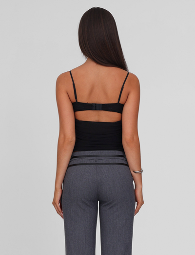 A woman with long, straight brown hair faces away, wearing the TIGERMIST KALLIE TOP - BLACK with adjustable straps and gray pants. A bracelet adorns her left arm, and her bare back is visible against a plain light background.