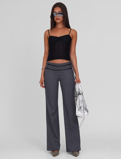 A woman wearing the TIGERMIST KALLIE TOP in black, gray wide-leg pants with an asymmetrical waistband, silver open-toe heels, sunglasses, and a shiny silver tote bag stands against a plain light gray background.