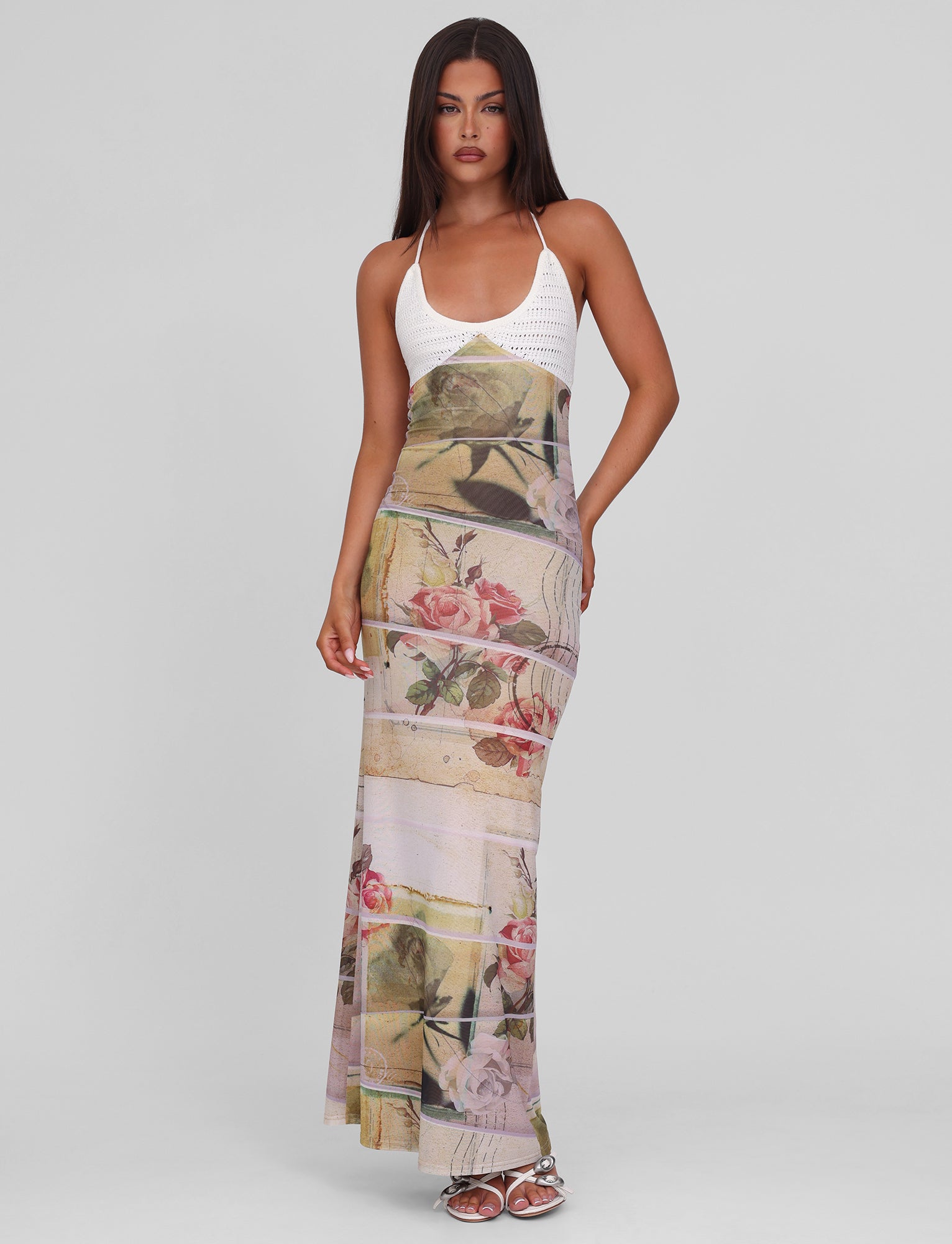 A woman with long dark hair and neutral makeup models the TIGERMIST TILLEY MAXI DRESS - ROSE POSTCARD, featuring crochet bust cups and a floral, abstract mesh skirt. She stands against a plain background wearing white sandals.
