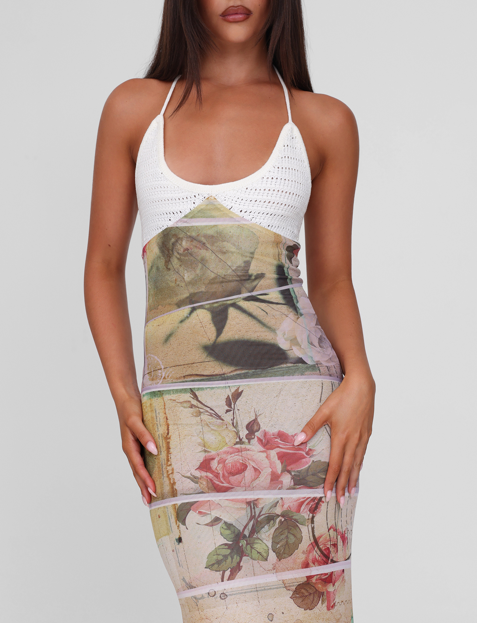 A woman wears the TIGERMIST TILLEY MAXI DRESS - ROSE POSTCARD, a sleeveless white crochet halter maxi dress with a fitted pink and red rose print skirt, standing against a plain light gray background.