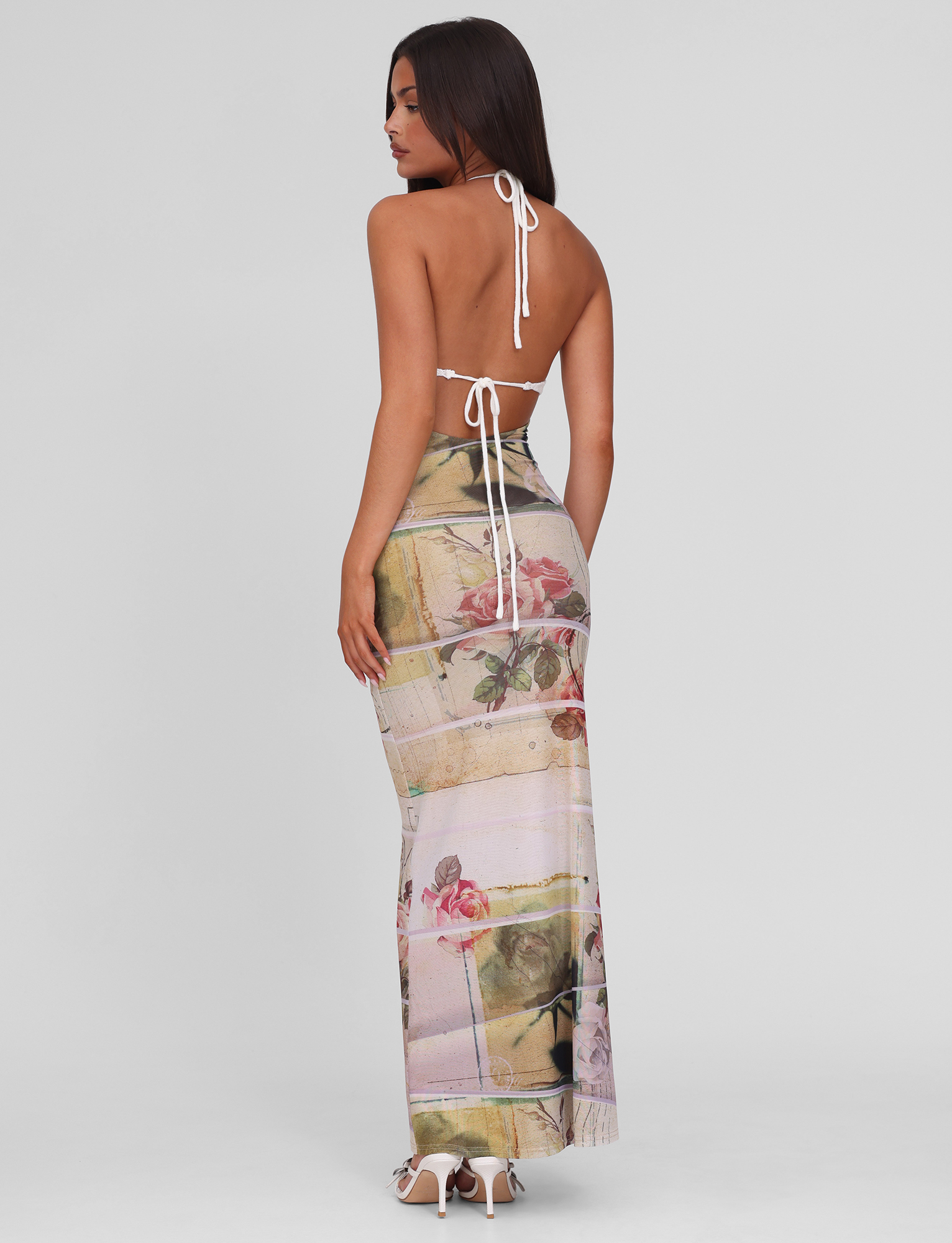 A woman stands with her back to the camera in the TIGERMIST TILLEY MAXI DRESS - ROSE POSTCARD, featuring an exclusive floral print and halter-tie backless design, paired with high heels against a plain light background.