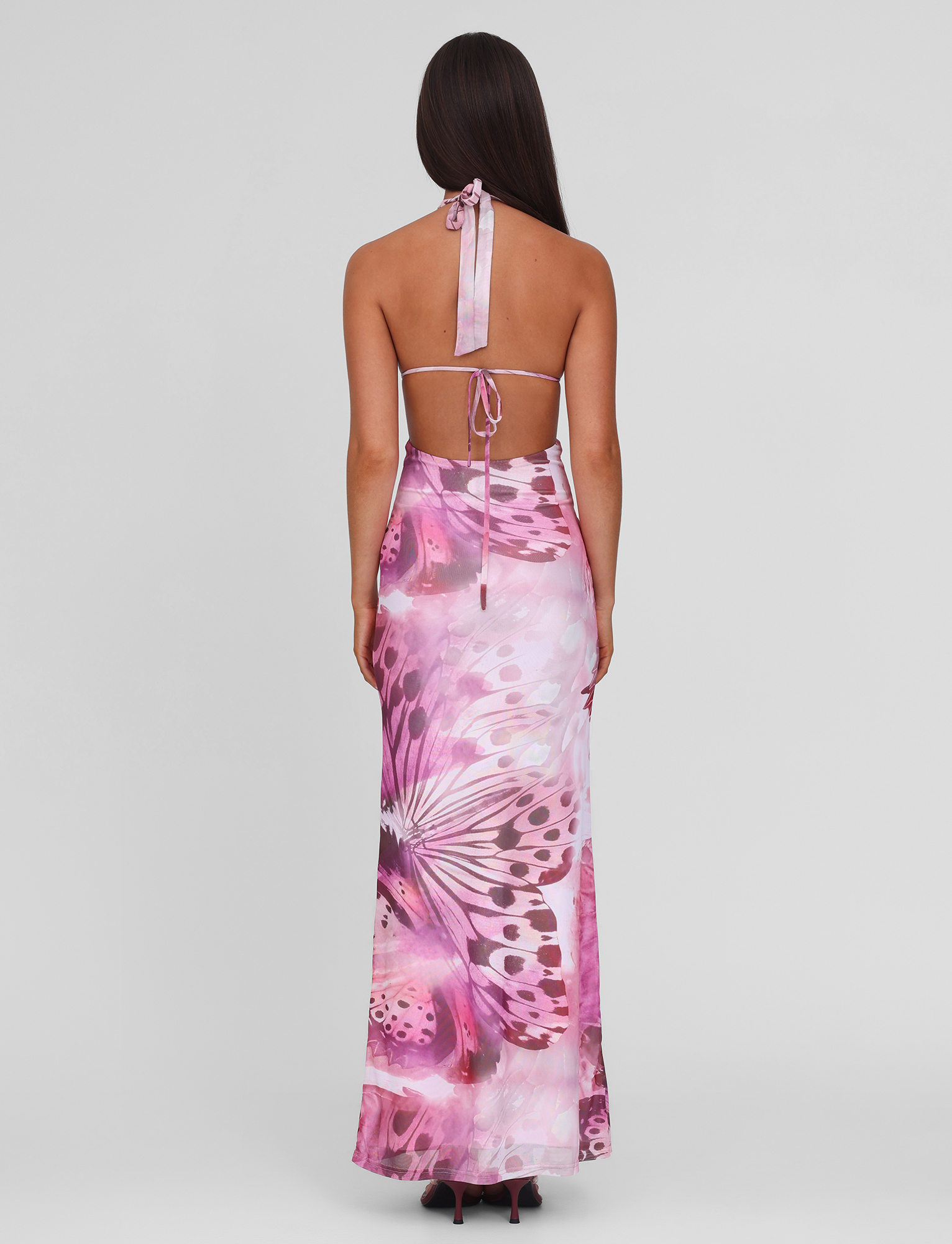 A woman with long brown hair models the TIGERMIST SOLEIL MAXI DRESS - MACRO BUTTERFLY, a mesh maxi dress with adjustable halter ties, standing against a plain light background with her back to the camera.