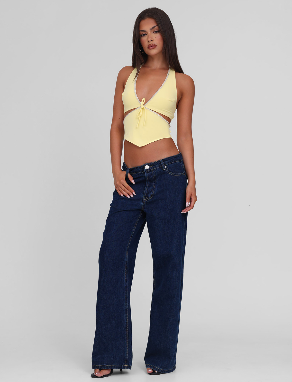 A woman with long brown hair and a neutral expression stands against a light gray background, wearing the TIGERMIST LIZZIE TOP in lemon and dark blue wide-leg jeans.