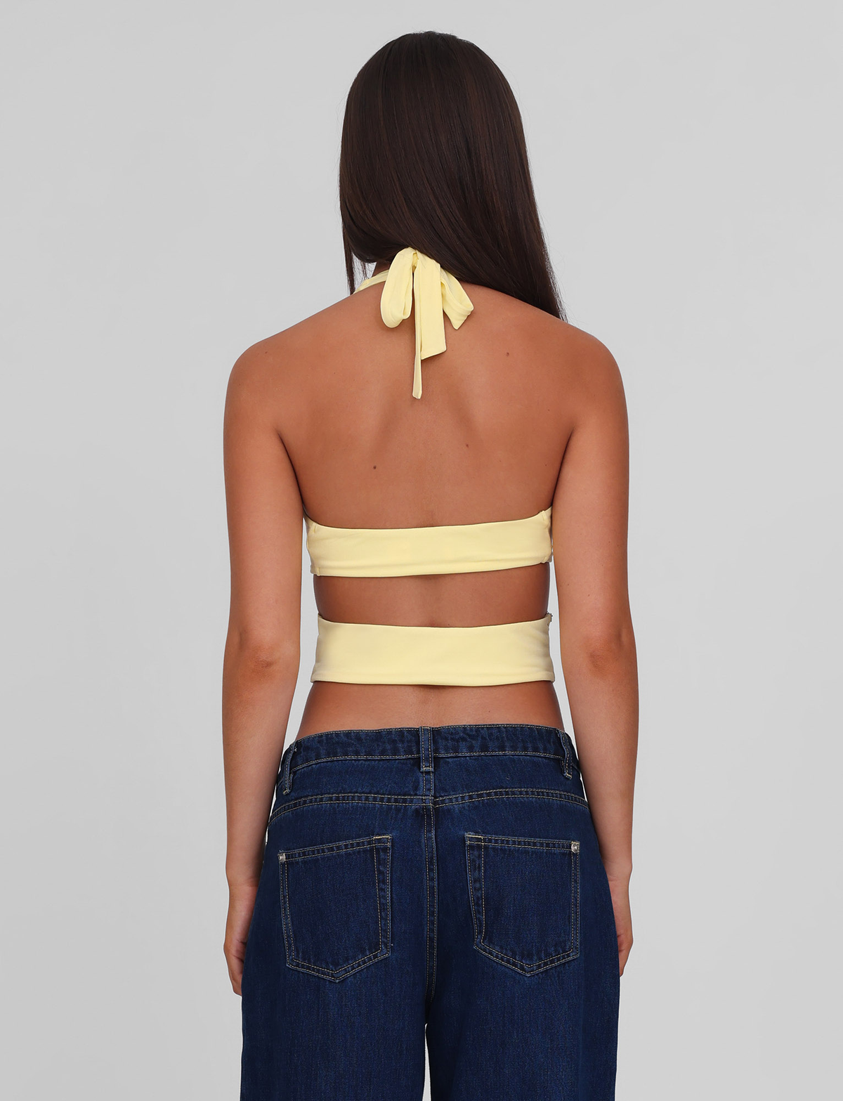 A woman with long brown hair, seen from behind, wears the TIGERMIST LIZZIE TOP in lemon (a yellow halter with cut-out details) and blue jeans against a plain gray background.