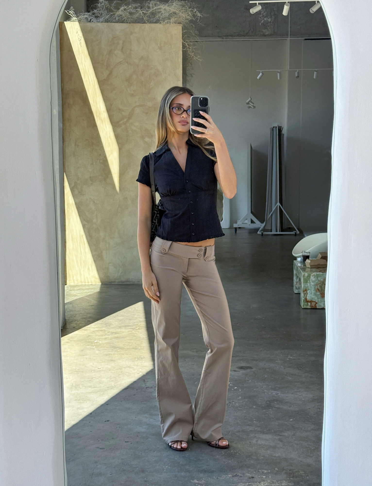 A woman wearing glasses, beige pants, and sandals takes a mirror selfie in a modern room, styled in the TIGERMIST VALERIE SHORT SLEEVE SHIRT - BLACK. Sunlight streams in, casting shadows on the concrete floor.