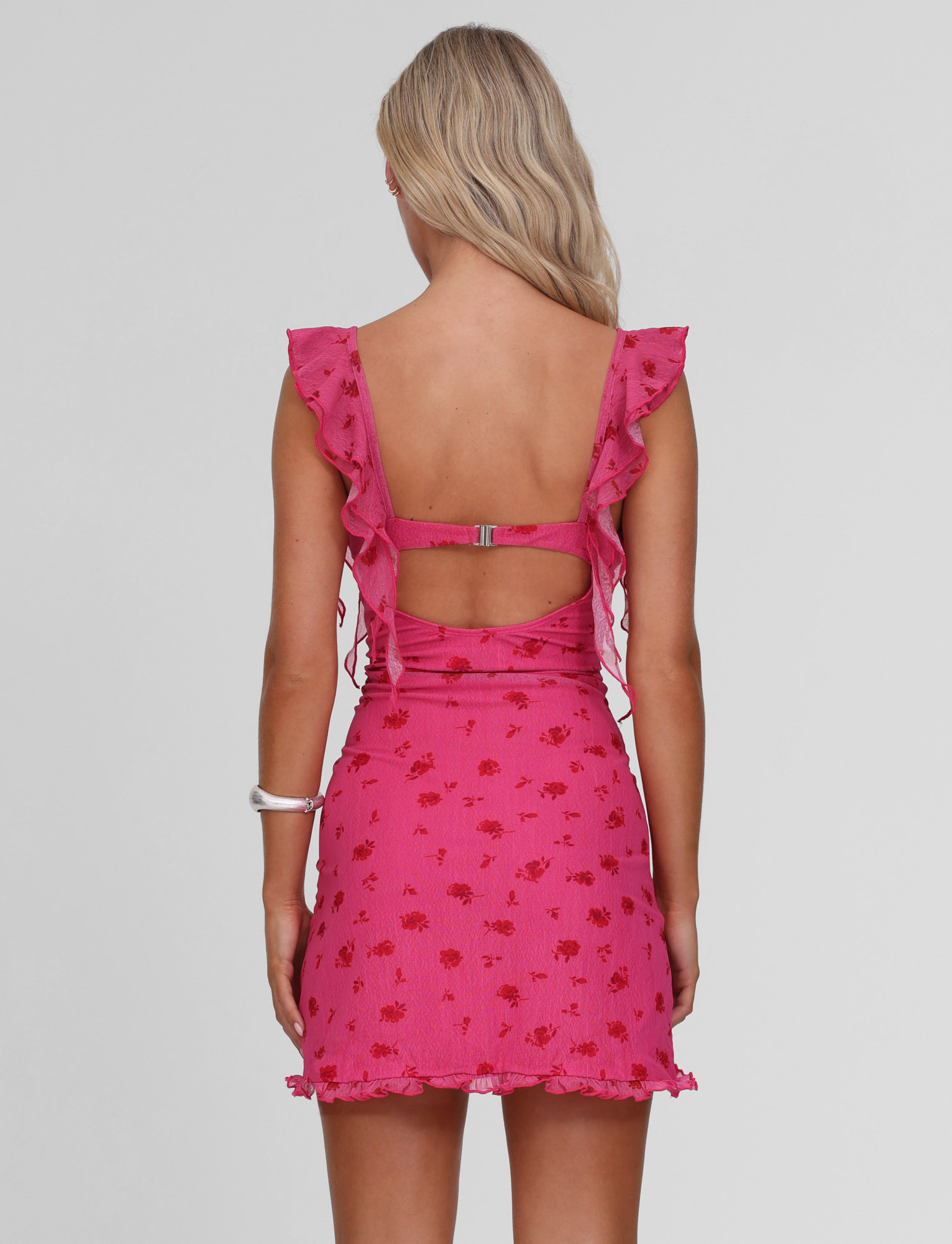 A woman with long blonde hair wears the TIGERMIST AMARIS MINI DRESS - DITSY FLORAL, featuring a pink V-neck, ruffle details, and a low open back, standing against a plain light gray background.