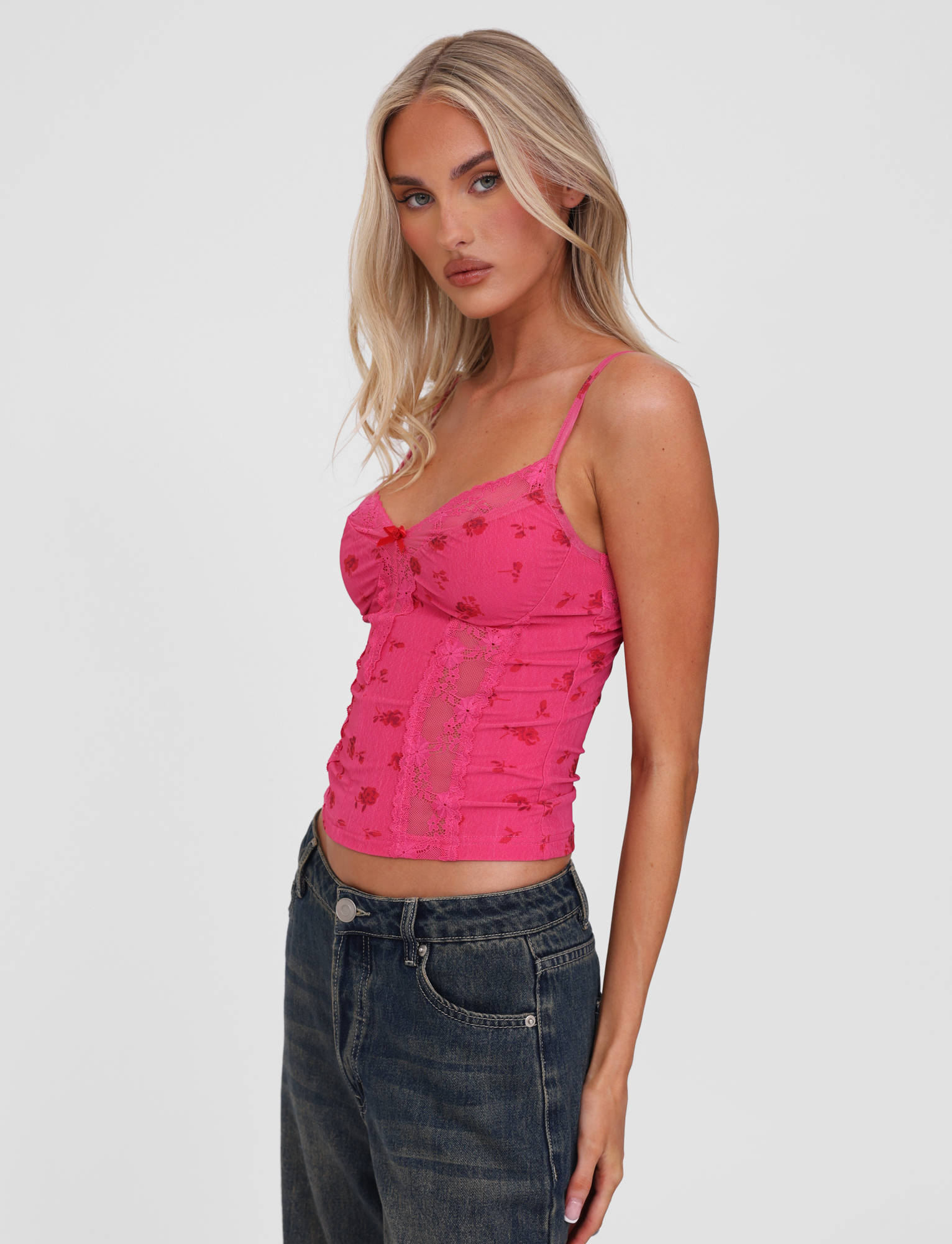 A woman with long blonde hair wears the TIGERMIST KHALIA TOP - DITSY FLORAL, a bright pink fitted sleeveless top with lace trim, paired with blue jeans, standing against a plain white background.