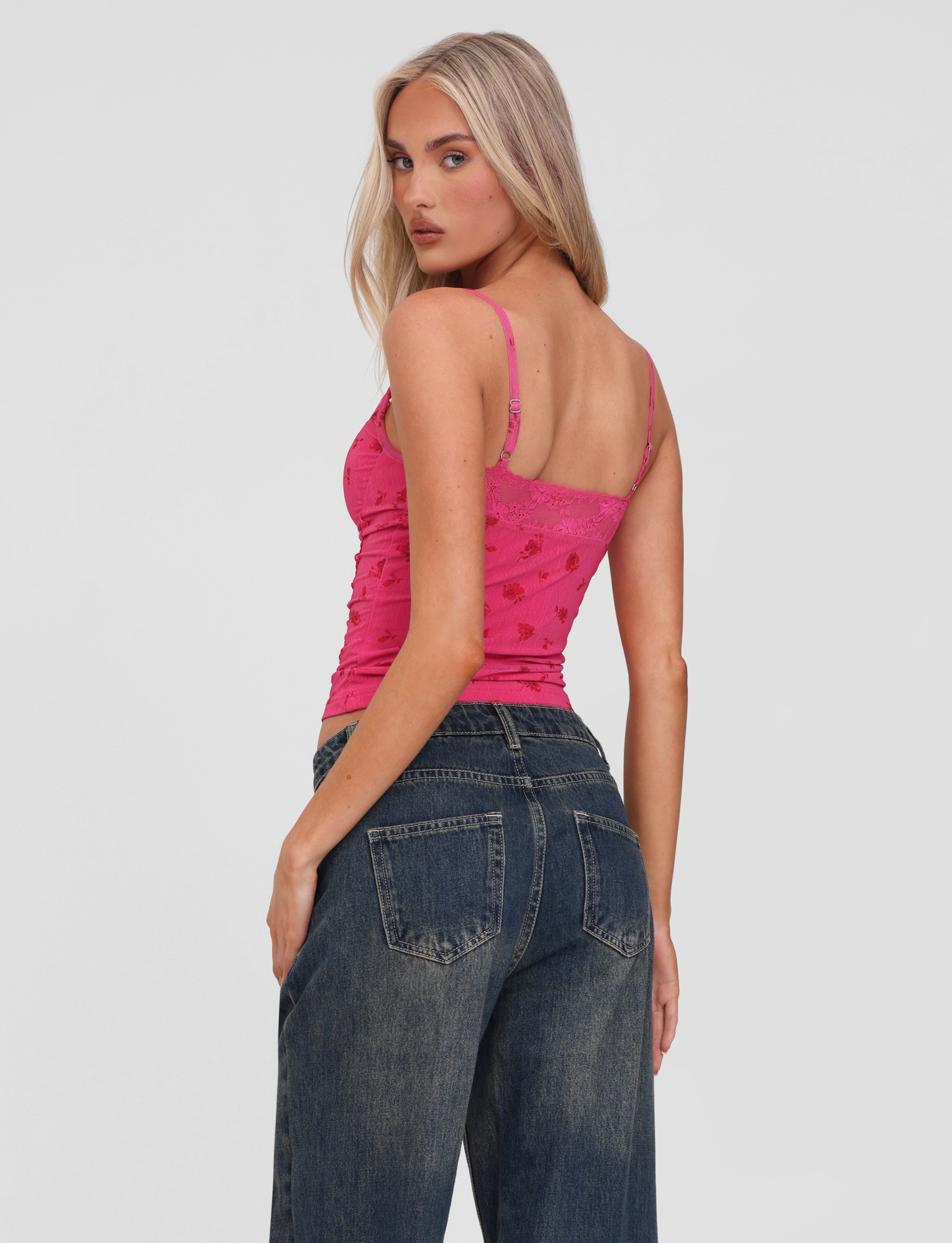 A woman with long blonde hair models the TIGERMIST KHALIA TOP - DITSY FLORAL, a pink sleeveless top with lace trim, paired with loose dark jeans. She poses with her back turned, looking over her shoulder against a plain white background.