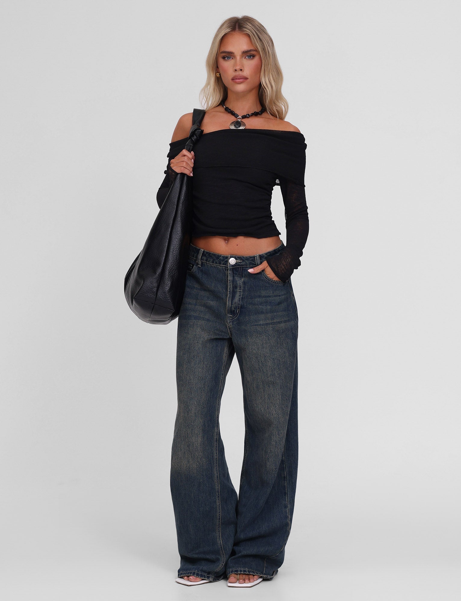A woman stands against a plain background wearing the TIGERMIST VERA TOP in black, wide-leg blue jeans, clear-heeled shoes, a black choker, and carrying a large black shoulder bag.