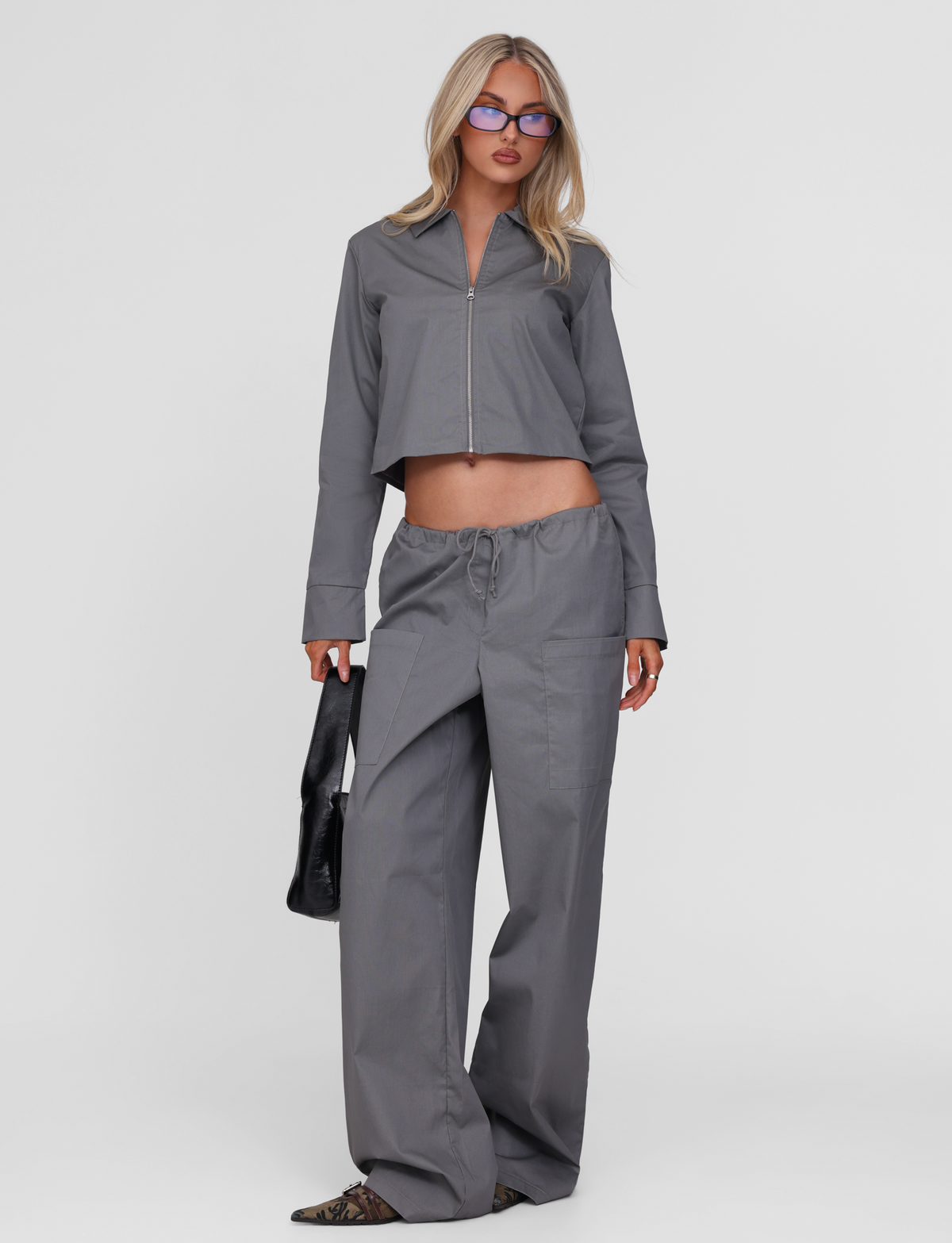 A woman stands against a plain white background wearing the TIGERMIST TYSHA JACKET in charcoal with matching wide-leg pants, pointed shoes, glasses, and holding a black handbag.