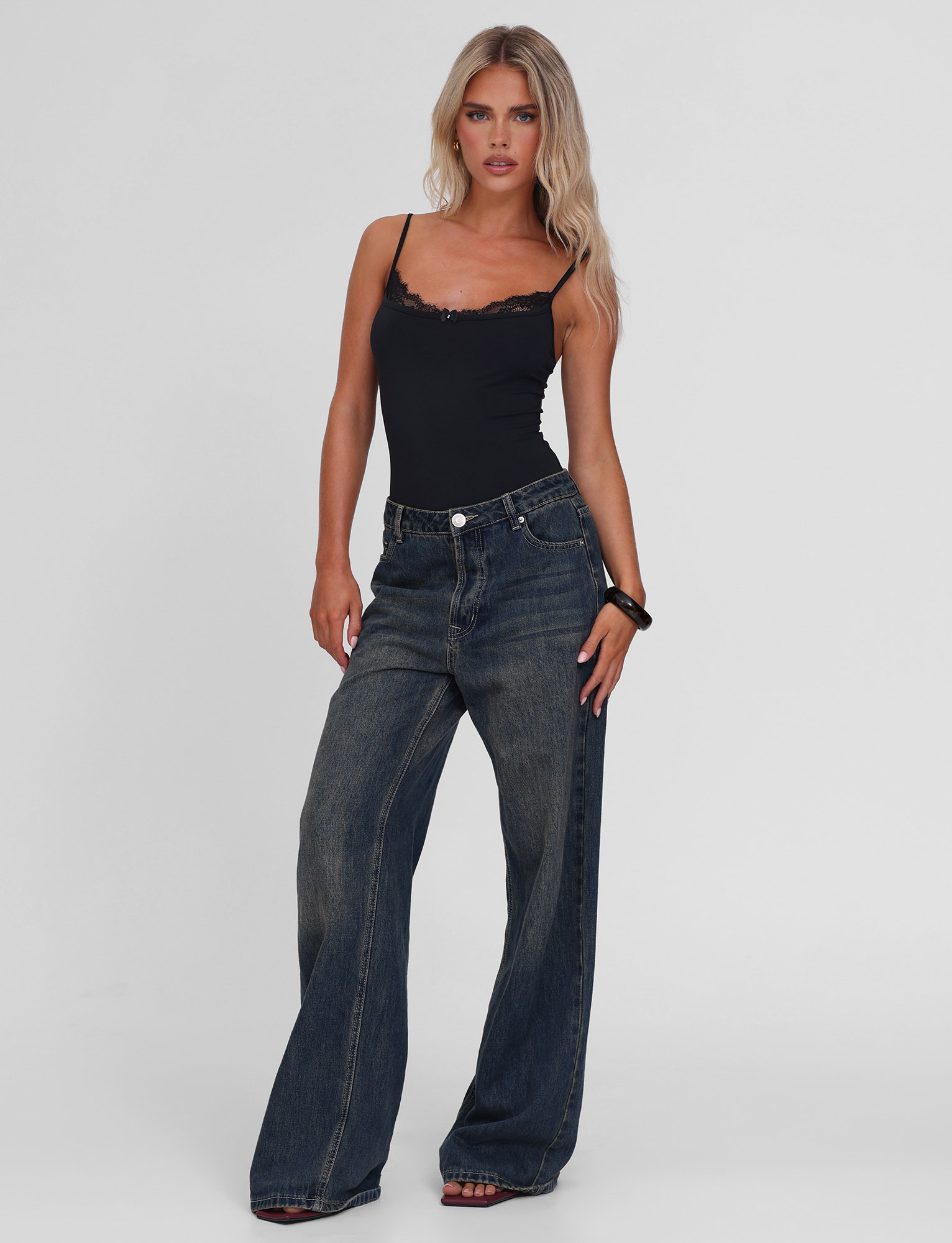 A woman with long blonde hair wears the TIGERMIST ASTRID BODYSUIT in black and wide-leg blue jeans. She stands against a white background, facing forward with one hand on her hip and the other resting by her side.