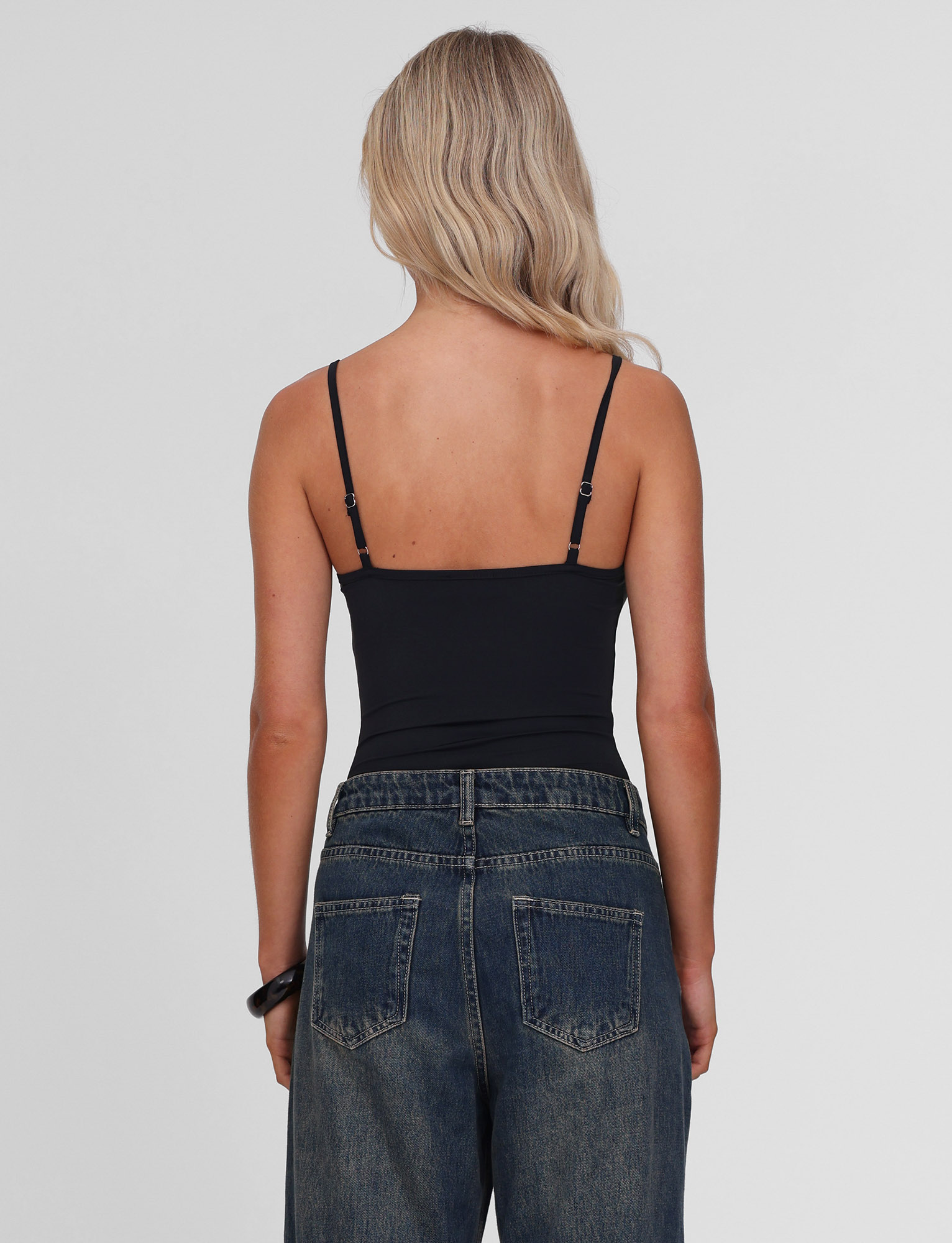 A person with long blonde hair is shown from the back, wearing the TIGERMIST ASTRID BODYSUIT - BLACK with adjustable straps and high-waisted blue jeans. The background is plain and light-colored.