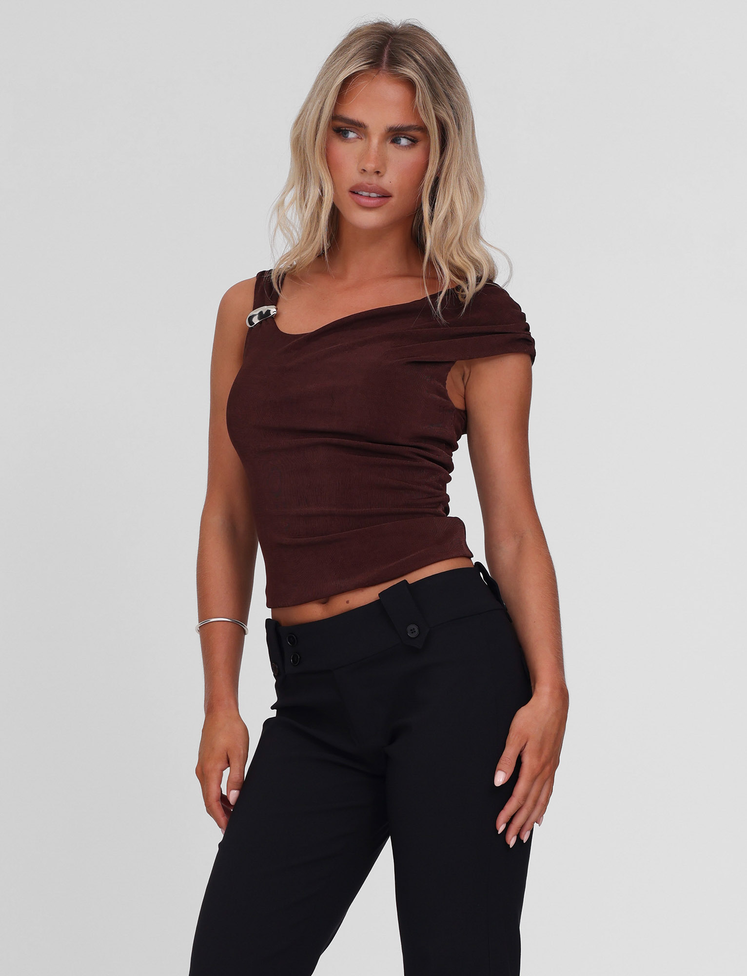 A woman with long blonde hair models the TIGERMIST CASSIAN TOP in chocolate, a slinky jersey one-shoulder style with metal detail, paired with black pants and posing against a plain light background.