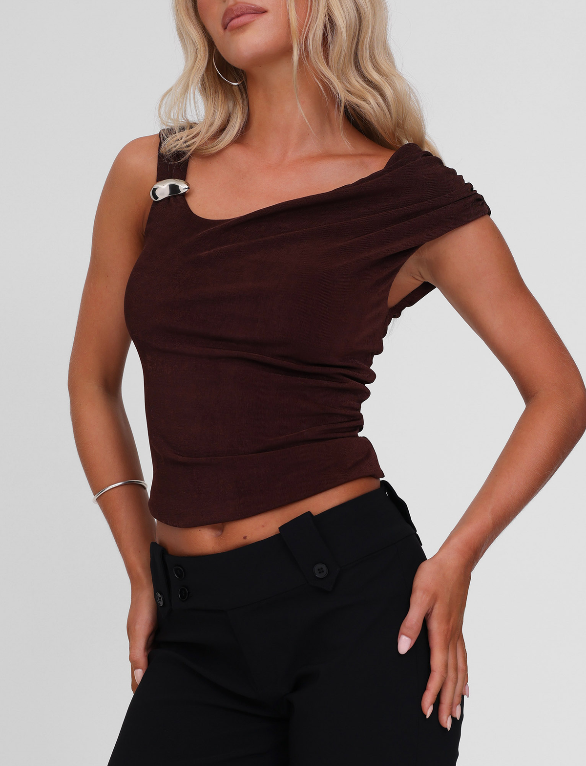 A woman stands with hands on hips against a plain background, wearing the TIGERMIST CASSIAN TOP in chocolate—a sleeveless tank featuring a metal buckle on one strap—paired with high-waisted black pants.