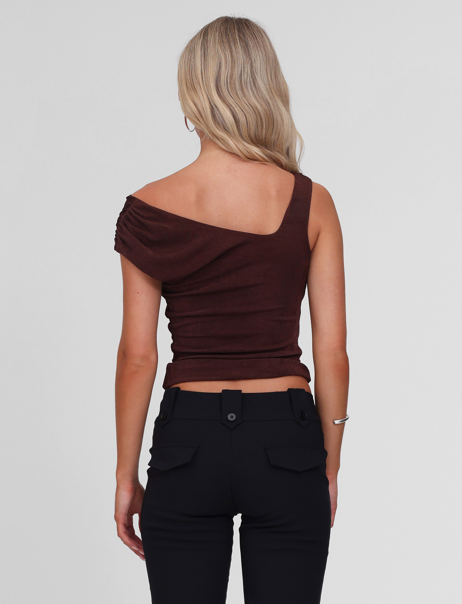 A woman with long blonde hair is shown from the back wearing the TIGERMIST CASSIAN TOP in chocolate and black pants with buttoned back pockets, standing against a plain white background.
