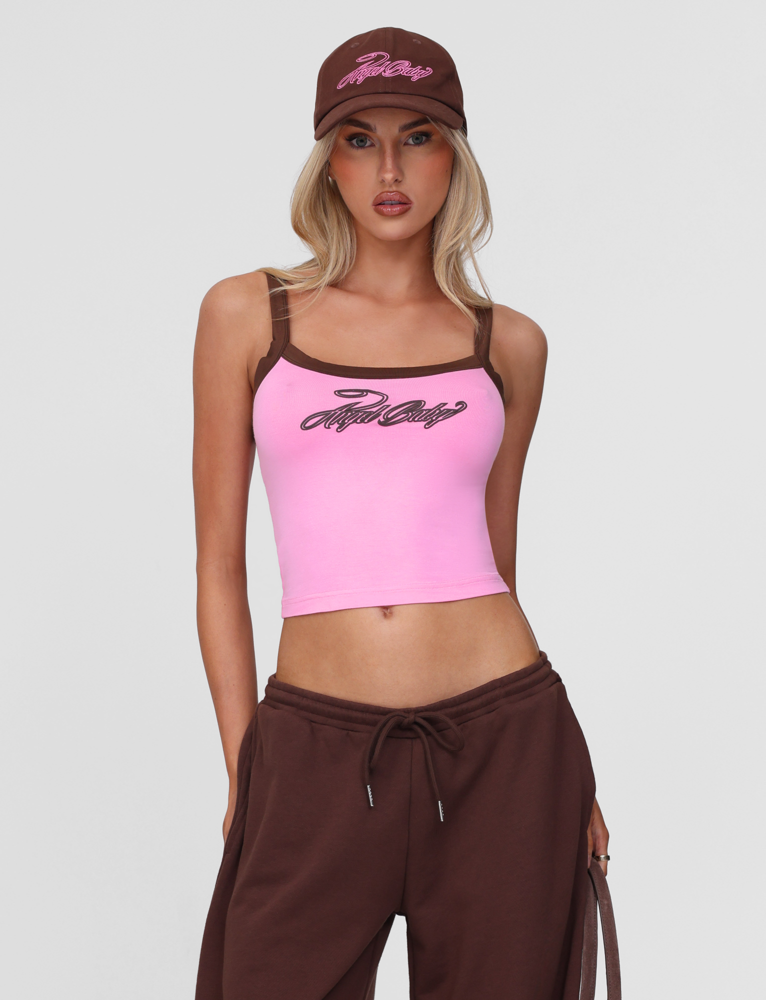 A woman stands against a plain light background wearing the TIGERMIST MERRYN TANK in pink, featuring script text and brown trim, paired with matching brown sweatpants and a brown baseball cap.