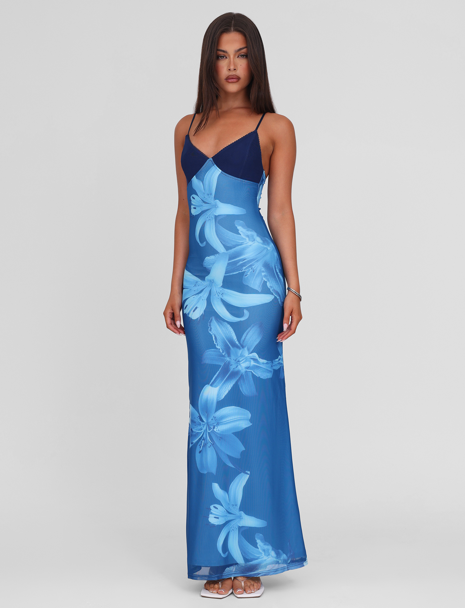 A woman with straight, dark hair stands against a plain background wearing the TIGERMIST ALAINA DRESS – BLUE ORCHID, a blue floral maxi dress with a V-neckline and adjustable spaghetti straps, paired with sandals. She has a neutral expression.