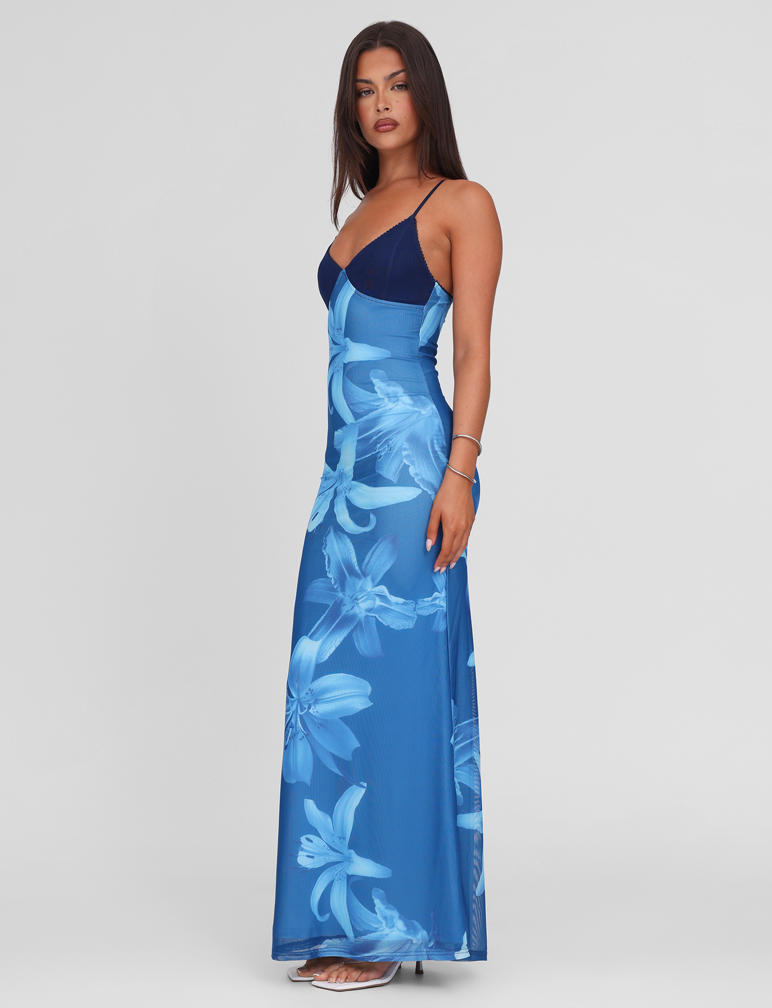 A woman poses against a white background, wearing the TIGERMIST ALAINA DRESS - BLUE ORCHID with a V-neckline and adjustable spaghetti straps. She has long straight hair and pairs the dress with open-toe heels.