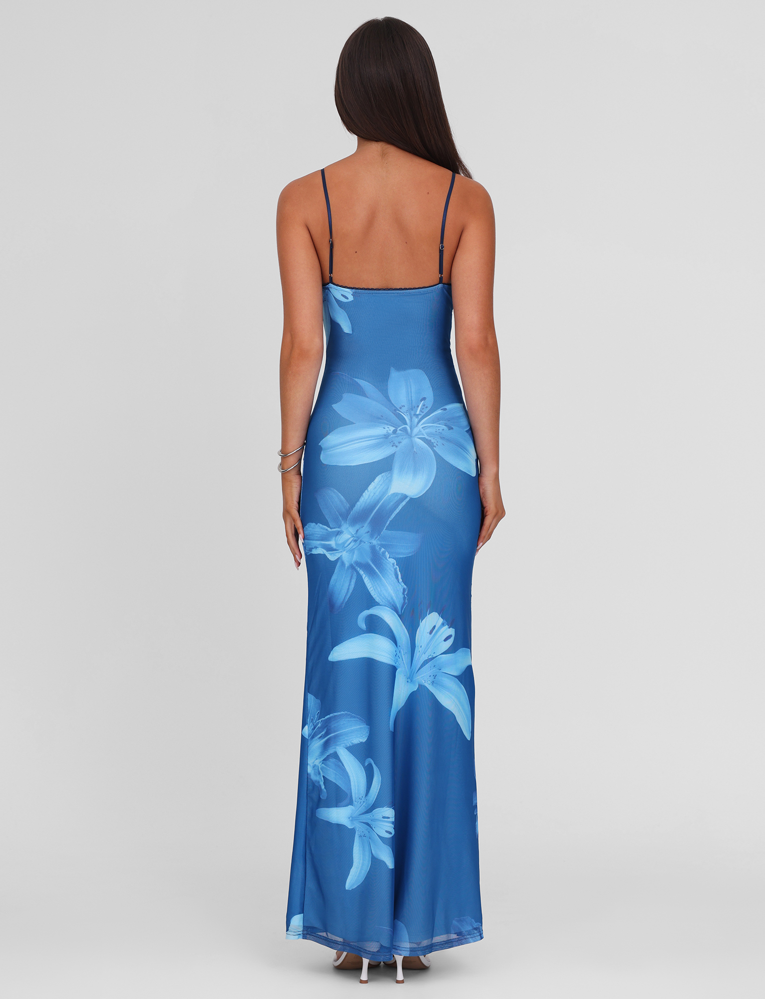 A woman with long dark hair, seen from the back, wears the TIGERMIST ALAINA DRESS - BLUE ORCHID, featuring adjustable spaghetti straps and bold light blue orchid prints, standing against a plain light background.