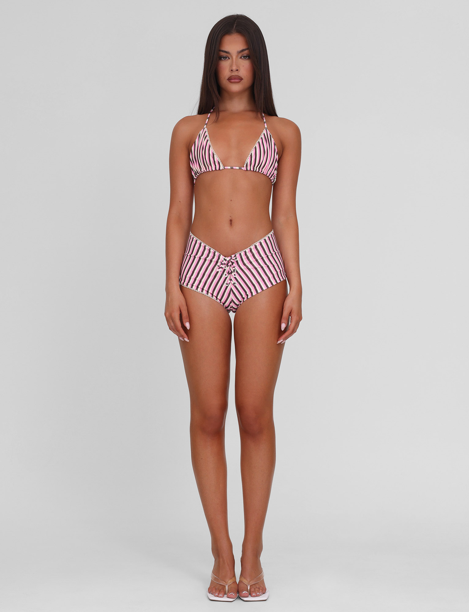 A woman with long dark hair stands facing forward in the TIGERMIST DAZZLE BIKINI TOP - STRIPE and clear sandals, set against a plain light gray background.