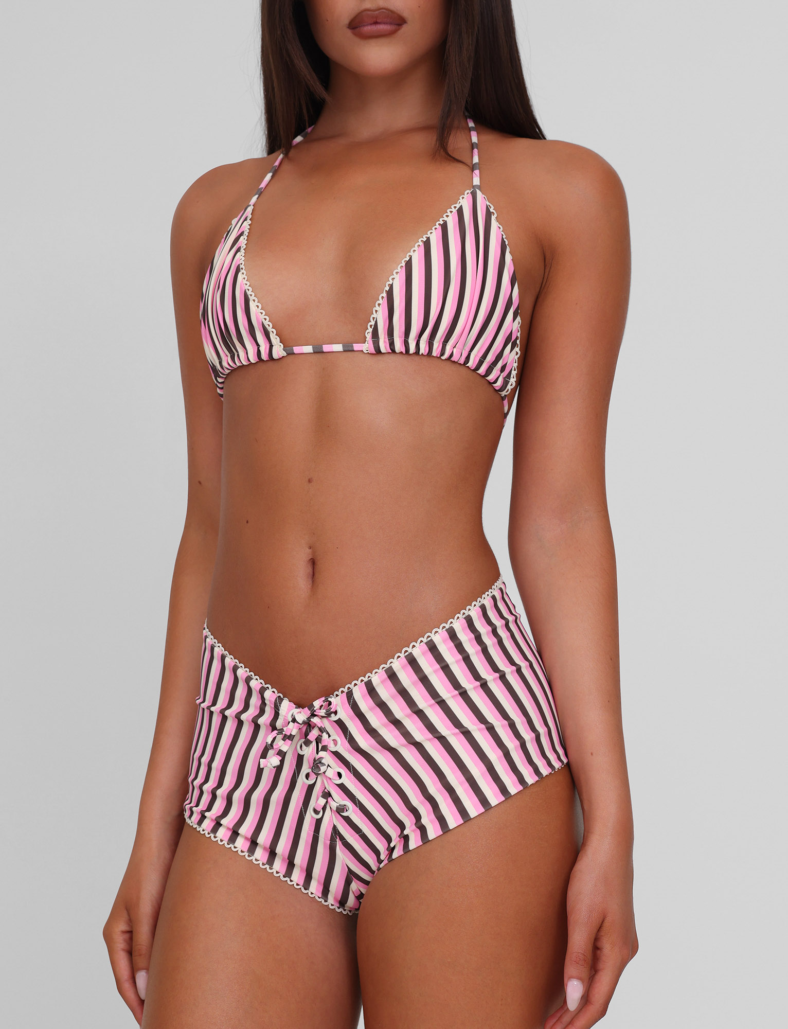 A woman models the TIGERMIST DAZZLE BIKINI TOP - STRIPE, featuring pink, black, and white stripes with scallop lace trim and lace-up high-waisted bottoms, against a plain light gray background.