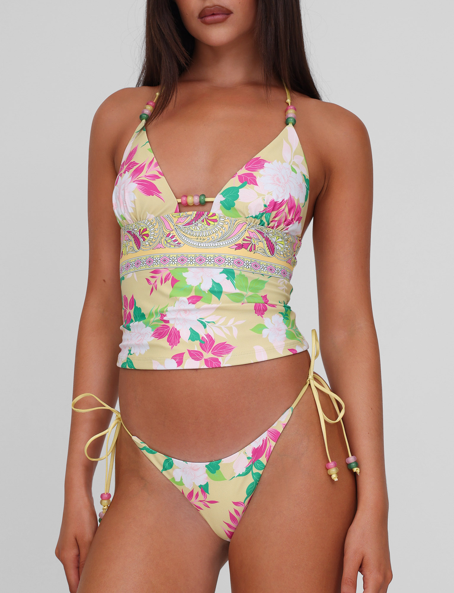 A woman wears the TIGERMIST FINLEA TANKINI TOP - PAISLEY BORDER with thin straps and matching tie-side bikini bottoms. The swimwear features a yellow paisley border print, and the background is plain light gray.