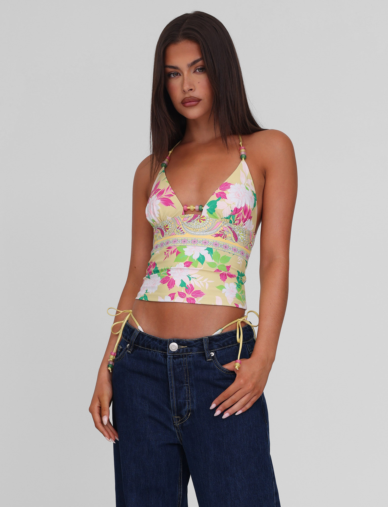 A woman with long straight brown hair wears the TIGERMIST FINLEA TANKINI TOP - PAISLEY BORDER and dark blue jeans, standing against a plain light gray background.