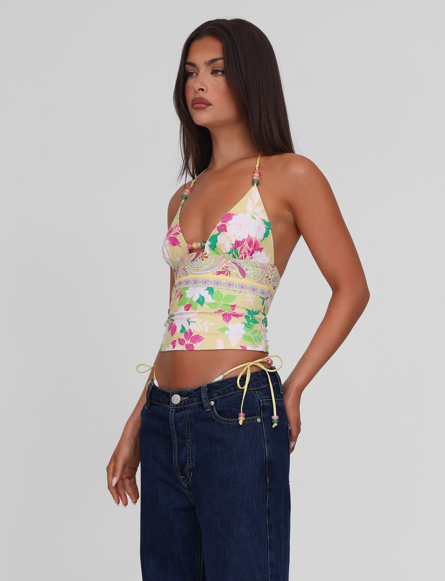 A woman with long dark hair poses against a light gray background wearing the TIGERMIST FINLEA TANKINI TOP in paisley border print and dark blue jeans.