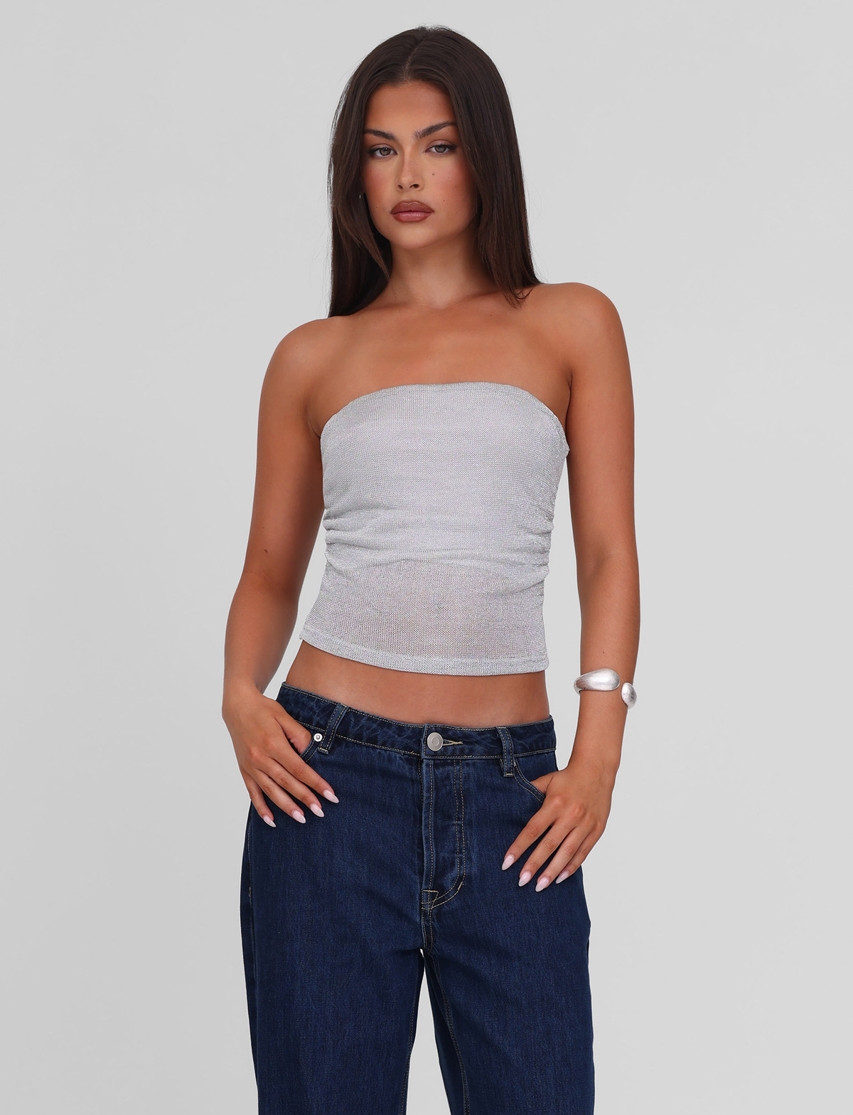 A woman with long dark hair wears the TIGERMIST HARRIETT TUBE TOP - SILVER, paired with dark blue jeans and a white bracelet. She stands against a plain light gray background with a neutral expression.