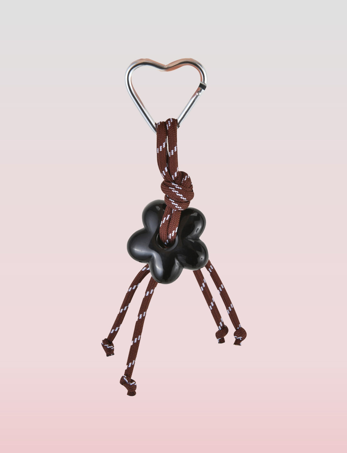 The TIGERMIST MIRIAM KEYCHAIN - CHOCOLATE boasts a charming heart-shaped metal clip as its centerpiece. It features a short brown cord with a black flower-shaped knot, four simply tied loose ends, all against a soothing soft pink gradient backdrop.