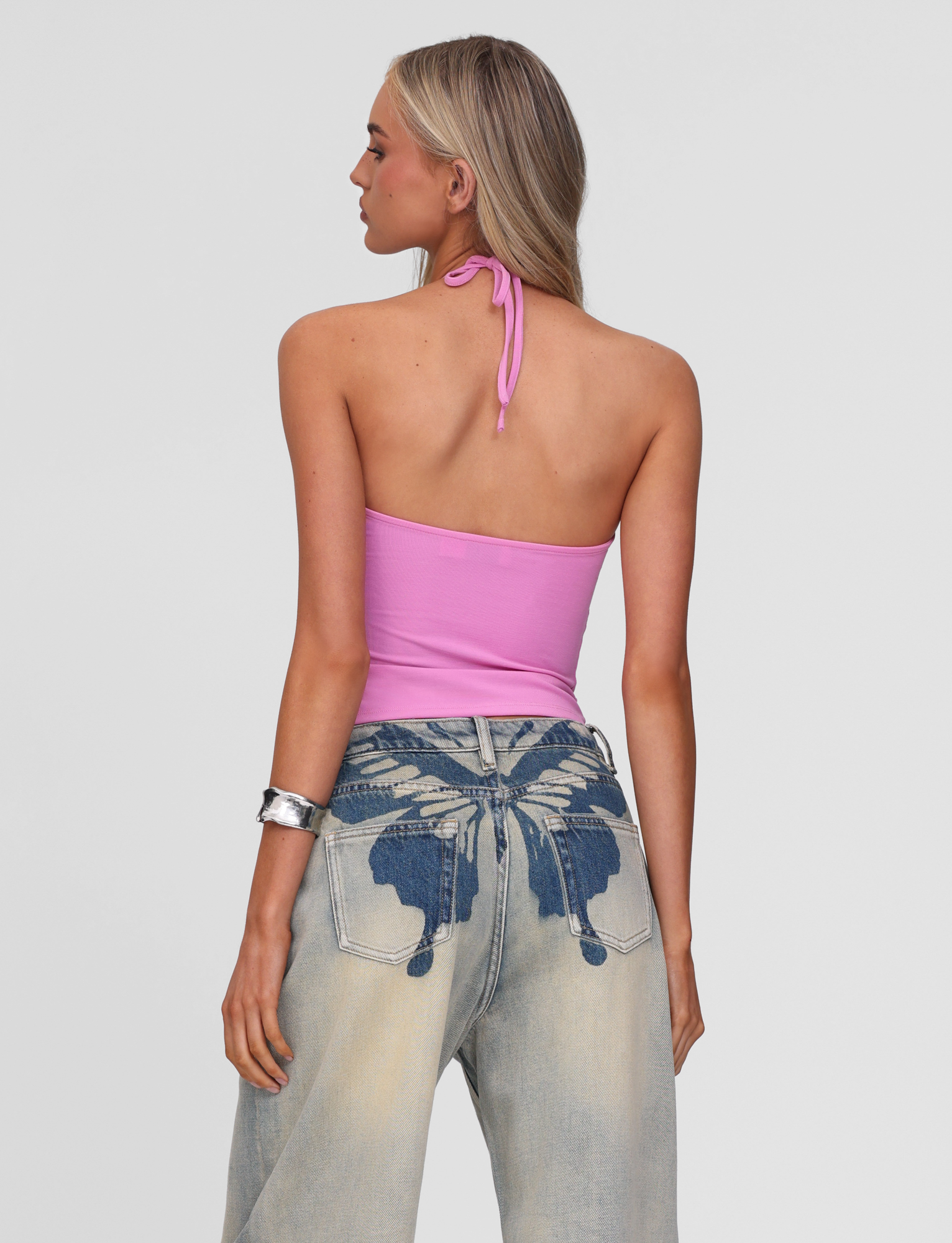 A woman with long blonde hair wears the TIGERMIST CELESTINE TOP in pink and light-wash jeans with embroidered butterfly trim on the back pockets, standing facing away against a plain background.