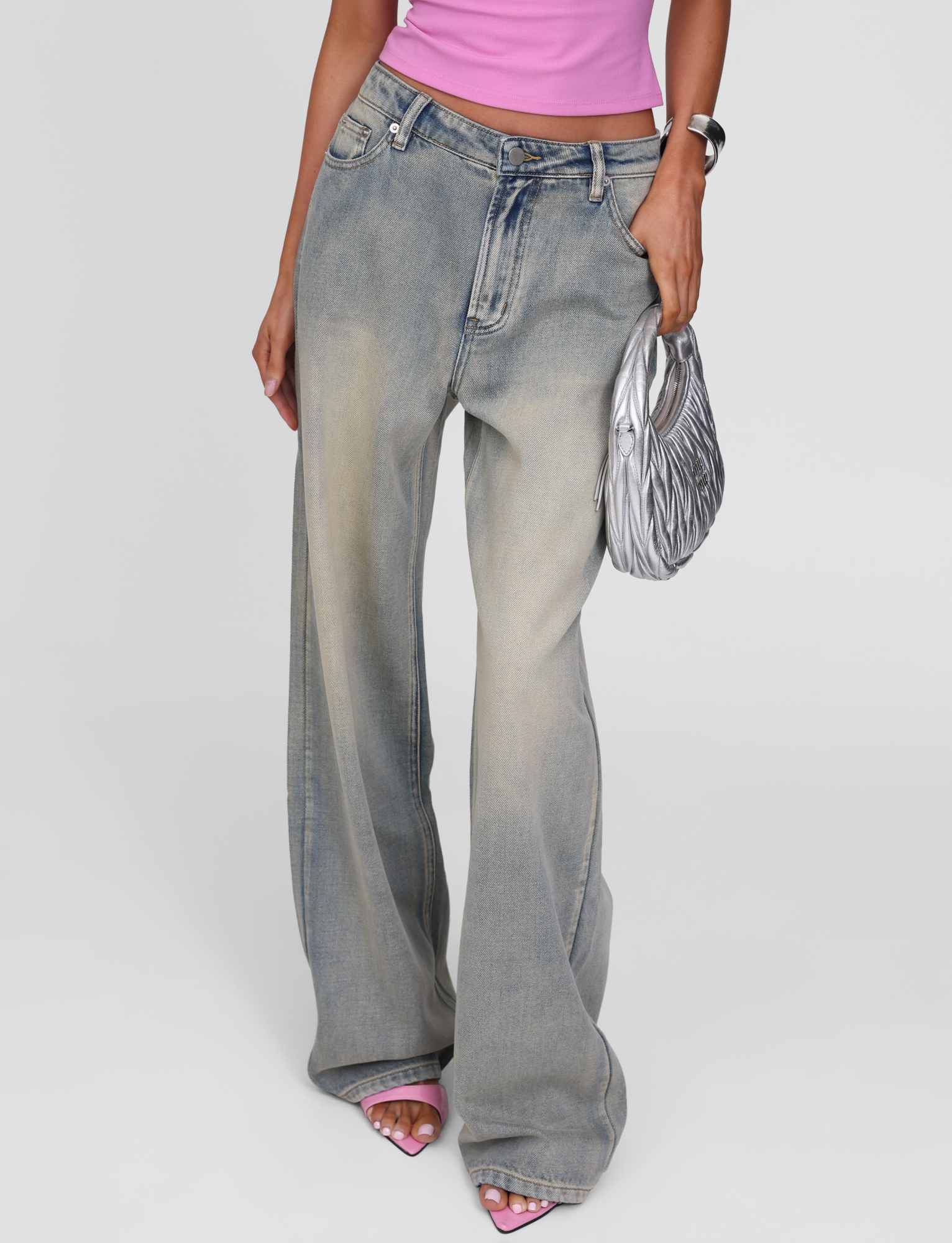 A person wearing TIGERMIST's ZANTHE JEAN in grey wash, a pink top, pink open-toe heels, a silver watch, and holding a shiny silver handbag. The background is plain white.