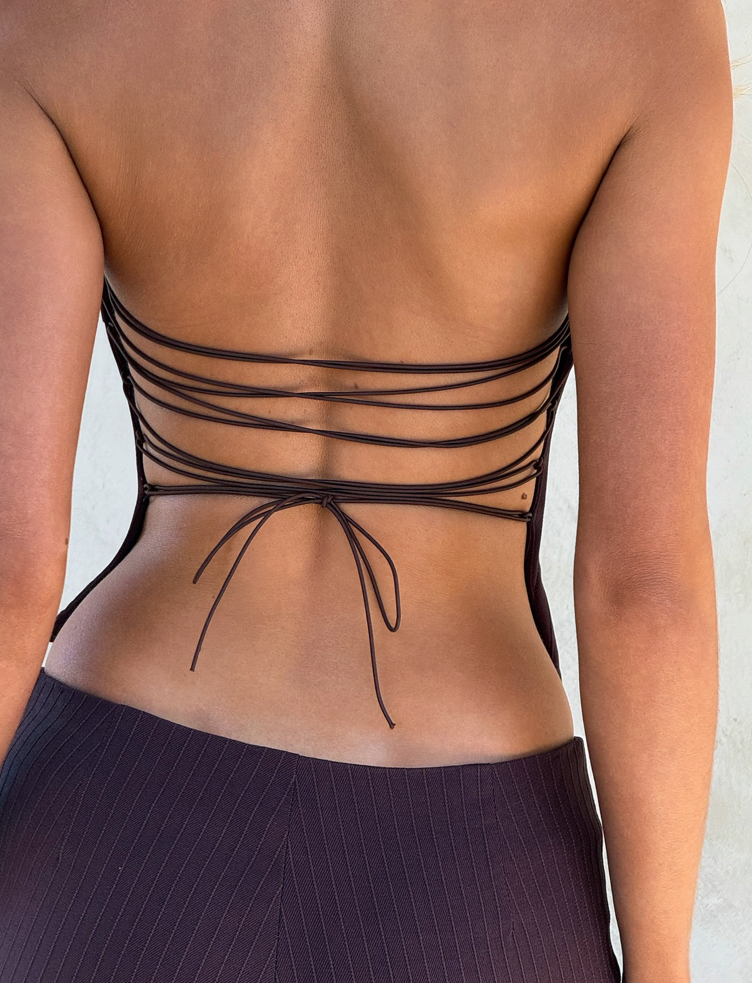 A close-up of a person wearing the TIGERMIST GWENDOLINE TOP in chocolate, featuring an open back with multiple thin crisscrossed straps tied in a bow, paired with dark textured pinstriped pants.