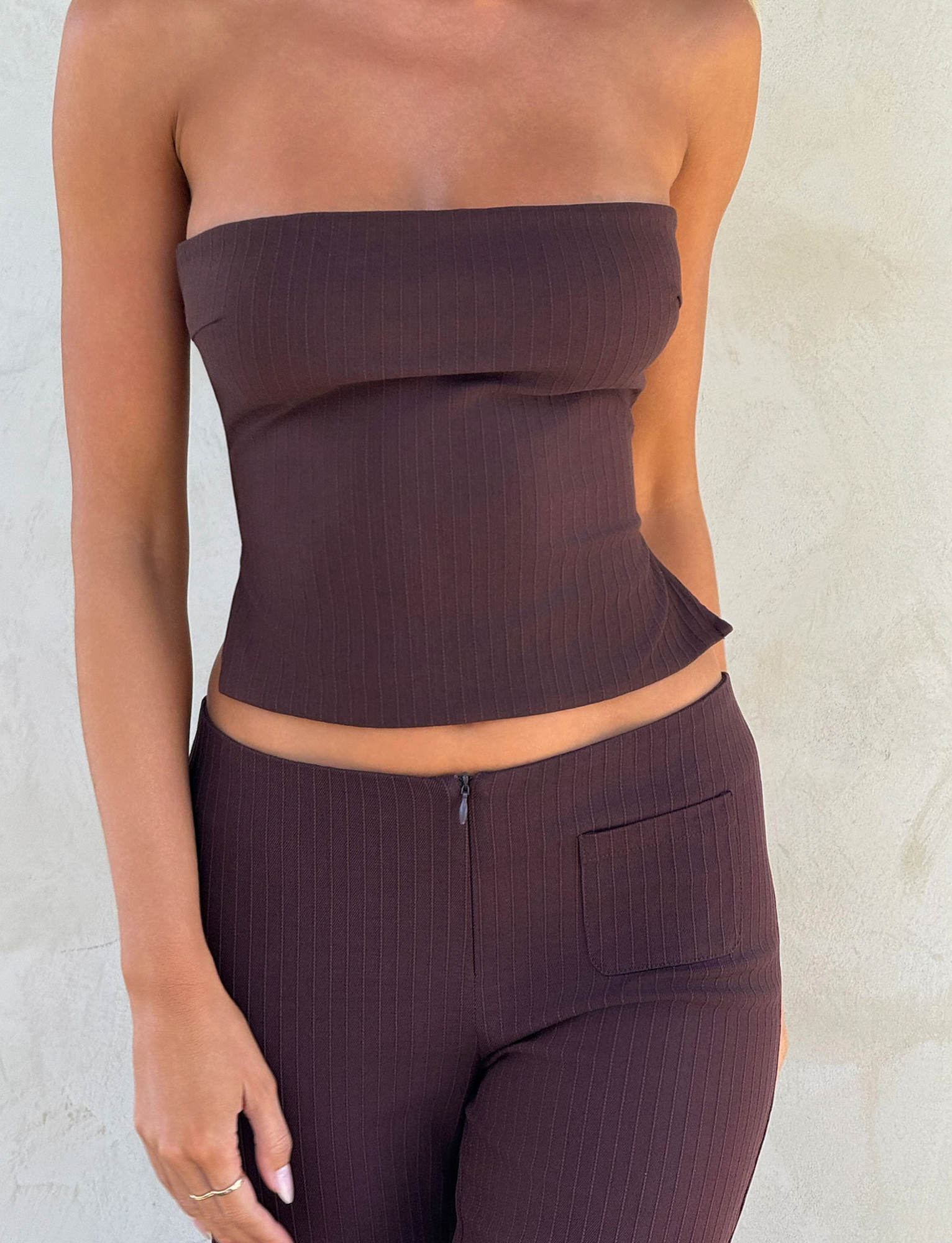 Wearing the TIGERMIST GWENDOLINE TOP - CHOCOLATE, this individual pairs the brown, pinstriped strapless piece with matching high-waisted pants, posing against a light textured wall in a cropped image that highlights the fitted look.