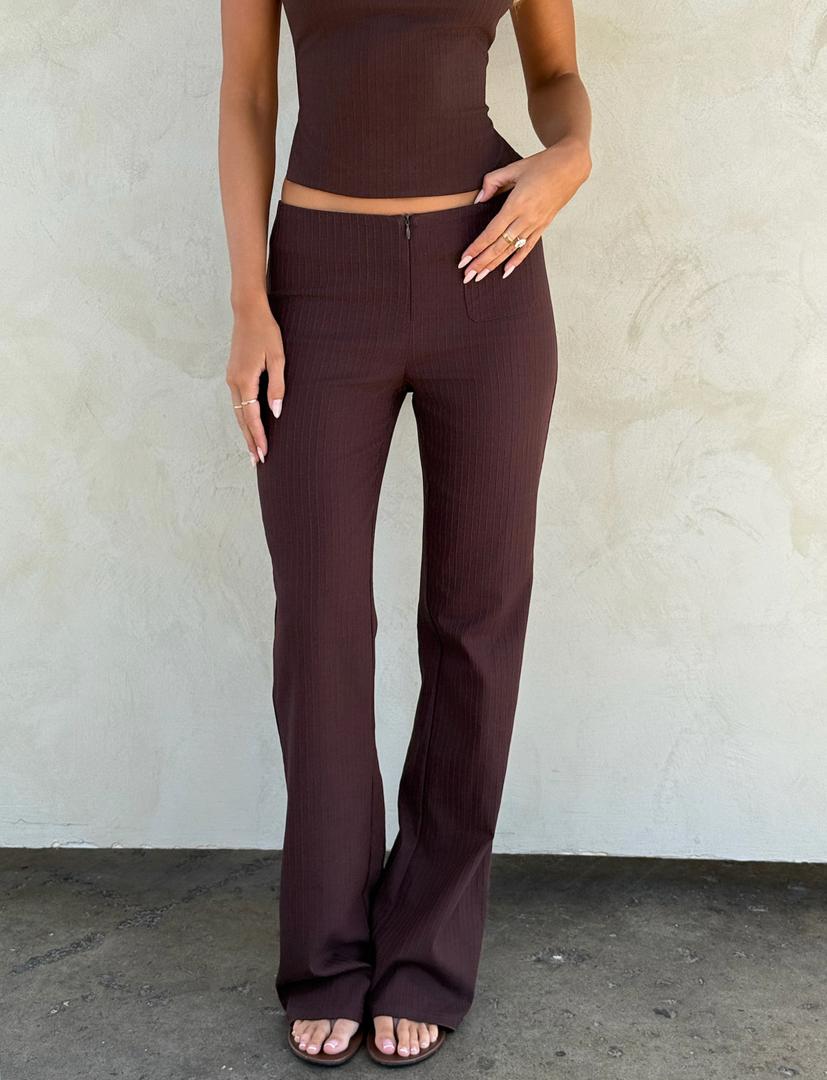 A woman stands barefoot against a textured wall, wearing TIGERMIST's GWENDOLINE PANT - CHOCOLATE, a high-waisted, flared-leg pant with a front zipper; her face is not visible.