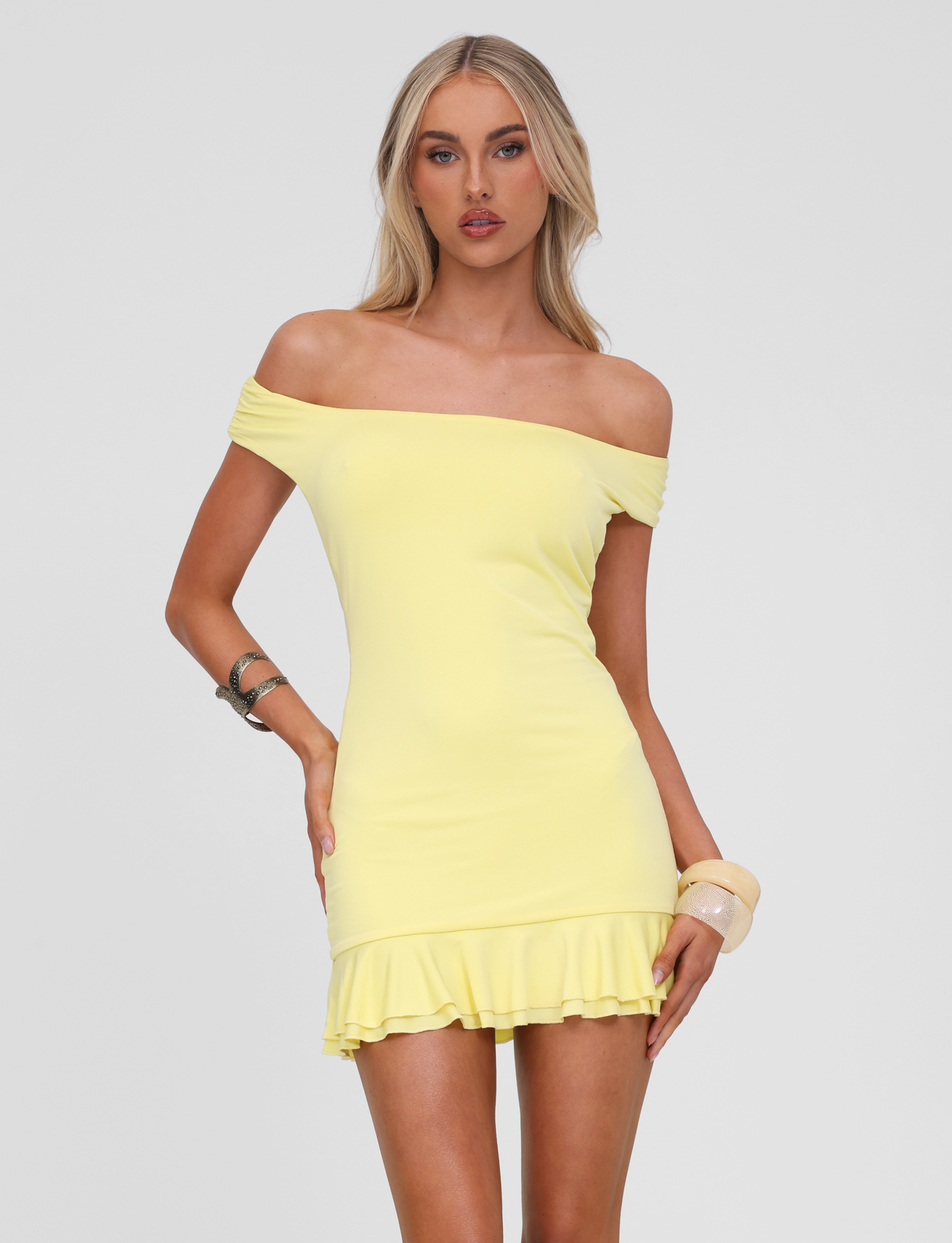 A woman with long blond hair models the TIGERMIST CLIA MINI DRESS in lemon, featuring an off-the-shoulder design and double frill hem, against a white background. She accessorizes with bangles on both wrists.