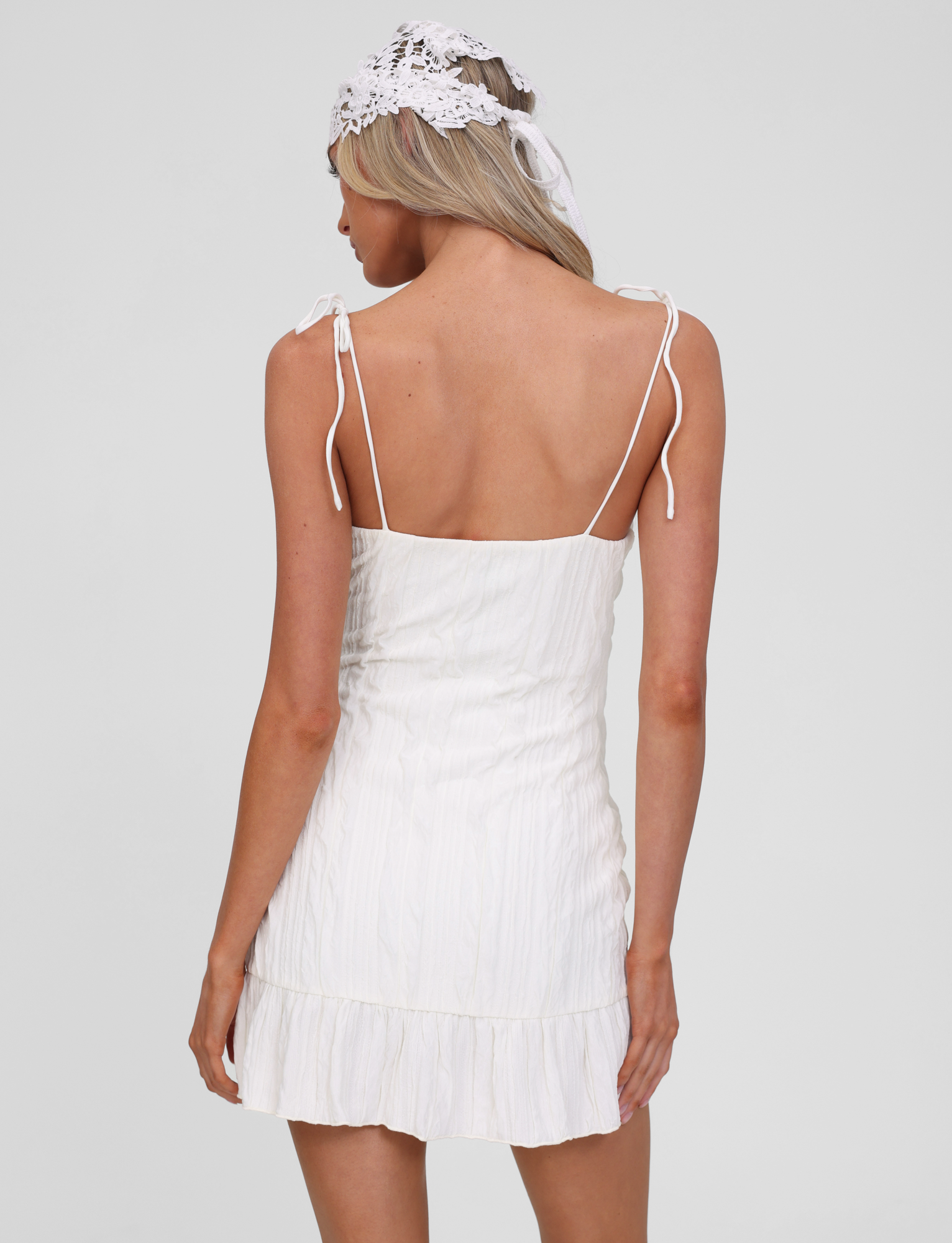A woman with long blonde hair models the TIGERMIST RANIA MINI DRESS in off white, featuring a frill hemline and low back, paired with a lace headpiece, standing against a plain light background.