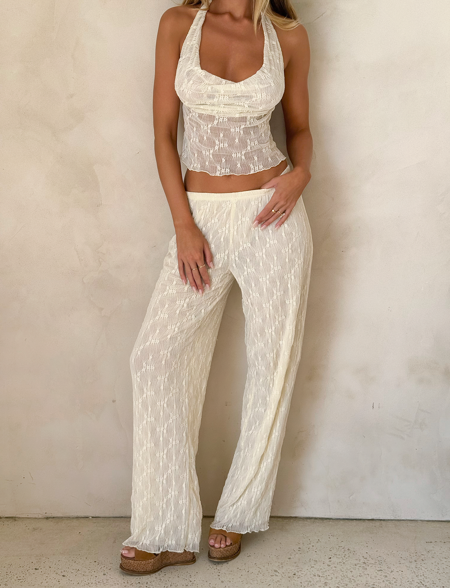 A woman poses against a light beige wall wearing TIGERMIST’s KATERINA PANT in ivory lace—wide-leg, floral, with an elastic waist—styled with a sleeveless white halter top and tan platform sandals. Her hands rest on her legs; her head is out of frame.
