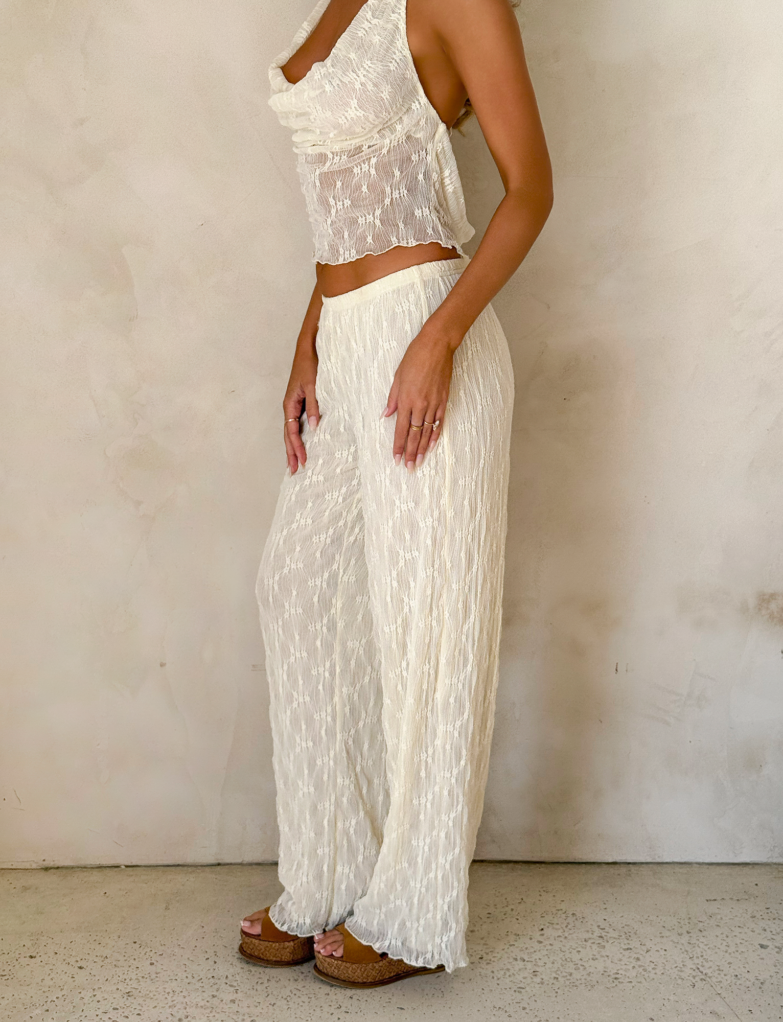 A woman stands against a light textured wall wearing the TIGERMIST KATERINA PANT - IVORY LACE with an elasticated waist, paired with a sheer white top and brown platform sandals. Only her body from shoulders to feet is visible.