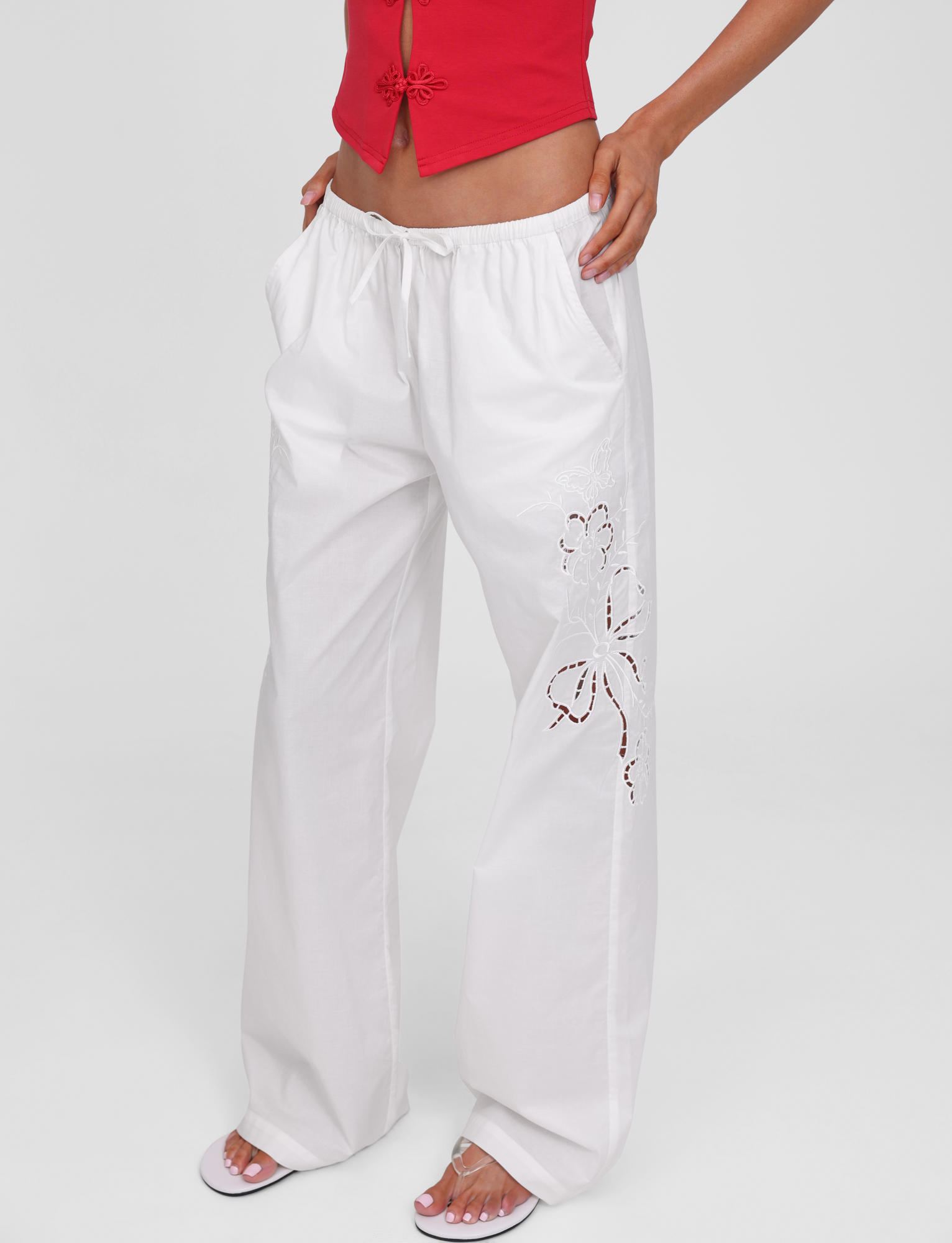 A person wears the TIGERMIST CRESSY PANT in white—wide leg, full length, cotton with one-leg embroidery—paired with a red sleeveless top and white sandals, standing against a plain white background.