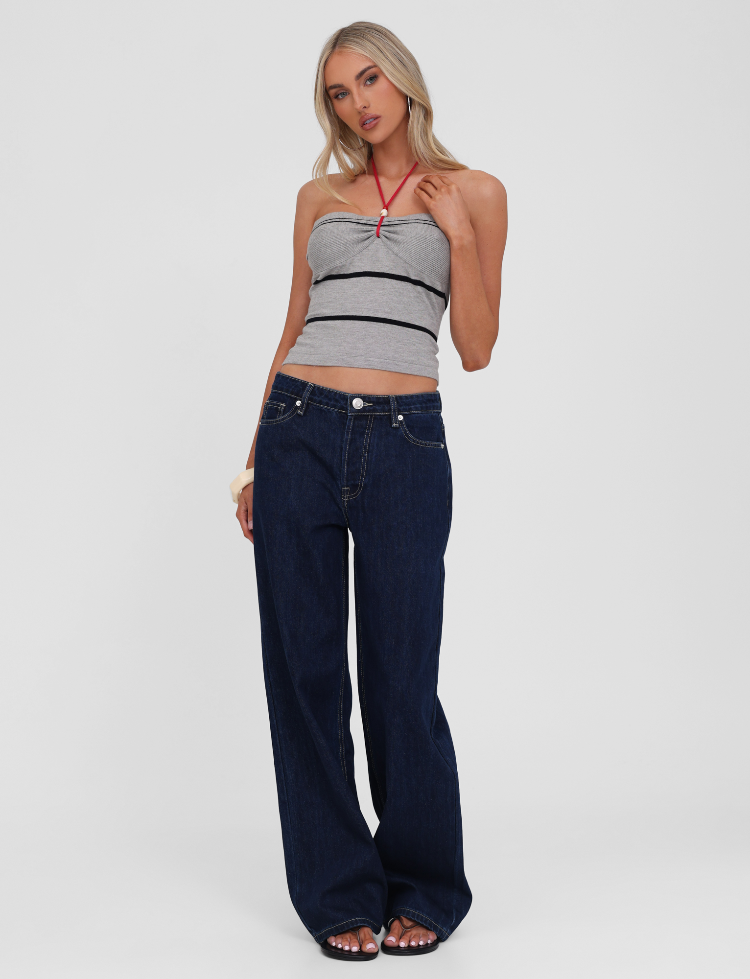 A woman with long blonde hair poses casually against a plain background, wearing the TIGERMIST NOUR TOP in grey stripe, dark blue wide-leg jeans, and open-toed shoes.