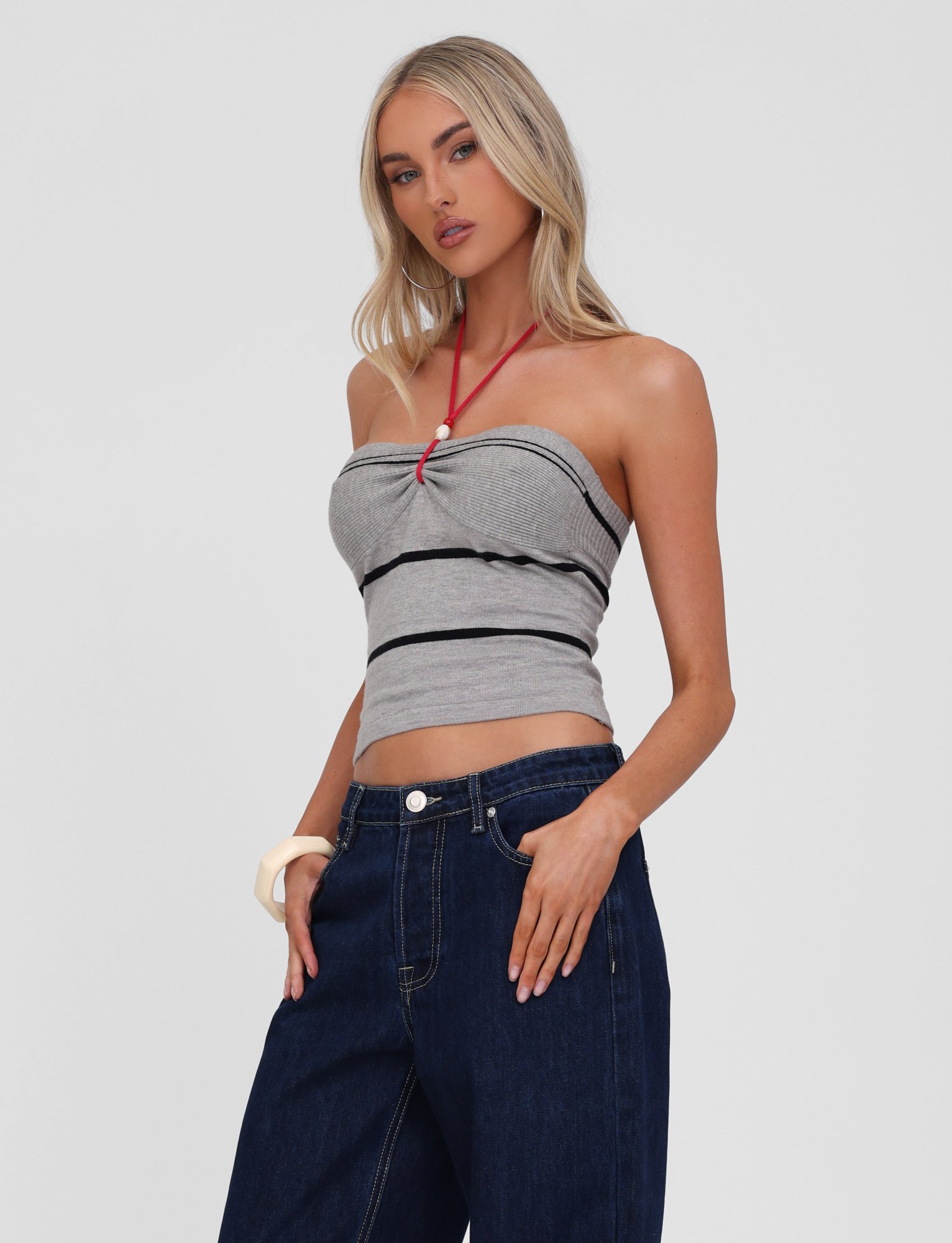 A woman with long blonde hair wears the TIGERMIST NOUR TOP in grey stripe and navy blue jeans, posing with one hand in her pocket against a plain light background.