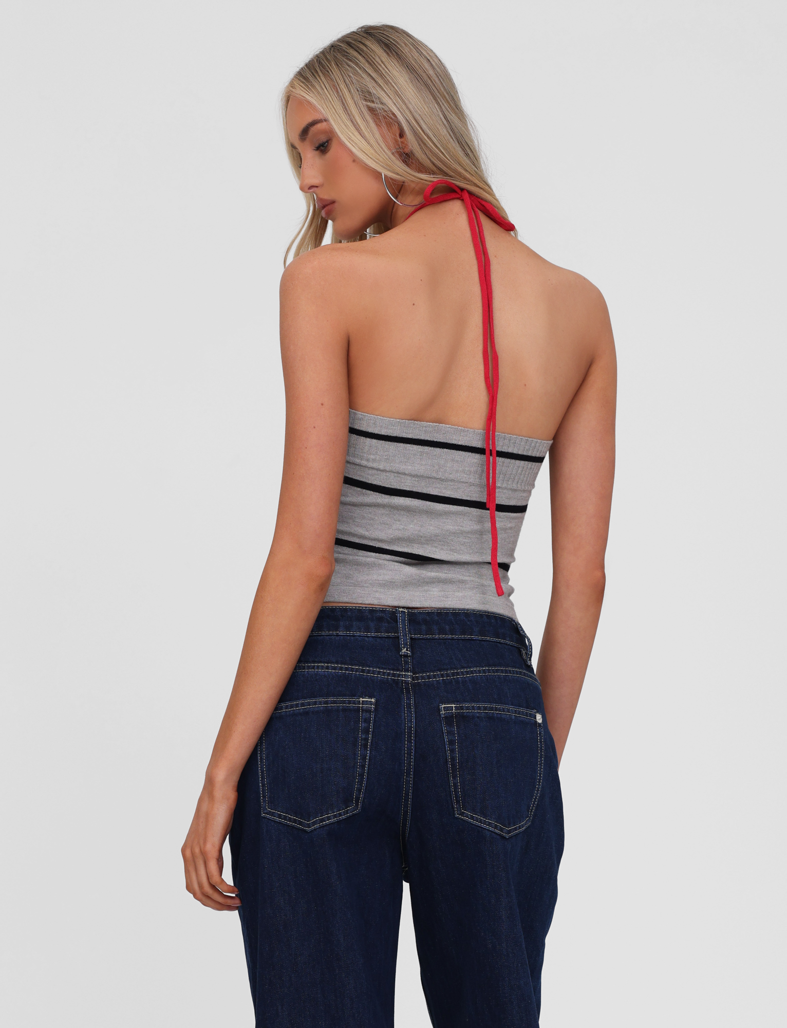 A woman with long blonde hair is seen from behind, wearing the TIGERMIST NOUR TOP in grey stripe and dark blue jeans, standing against a plain light background.