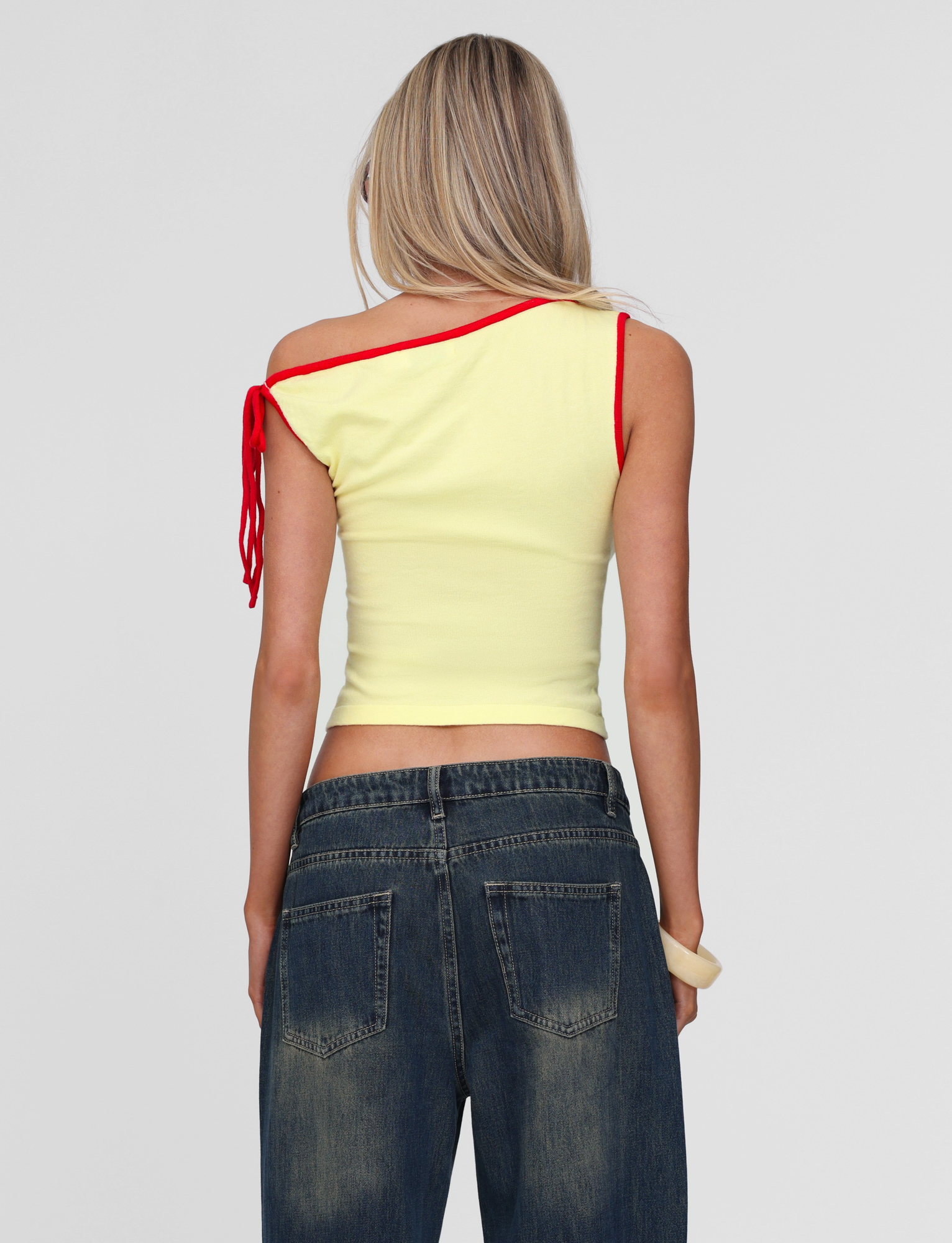 A woman with long blonde hair models the TIGERMIST ALIRA TOP in yellow, featuring red trim, paired with blue jeans. She stands with her back to the camera against a plain light gray background.